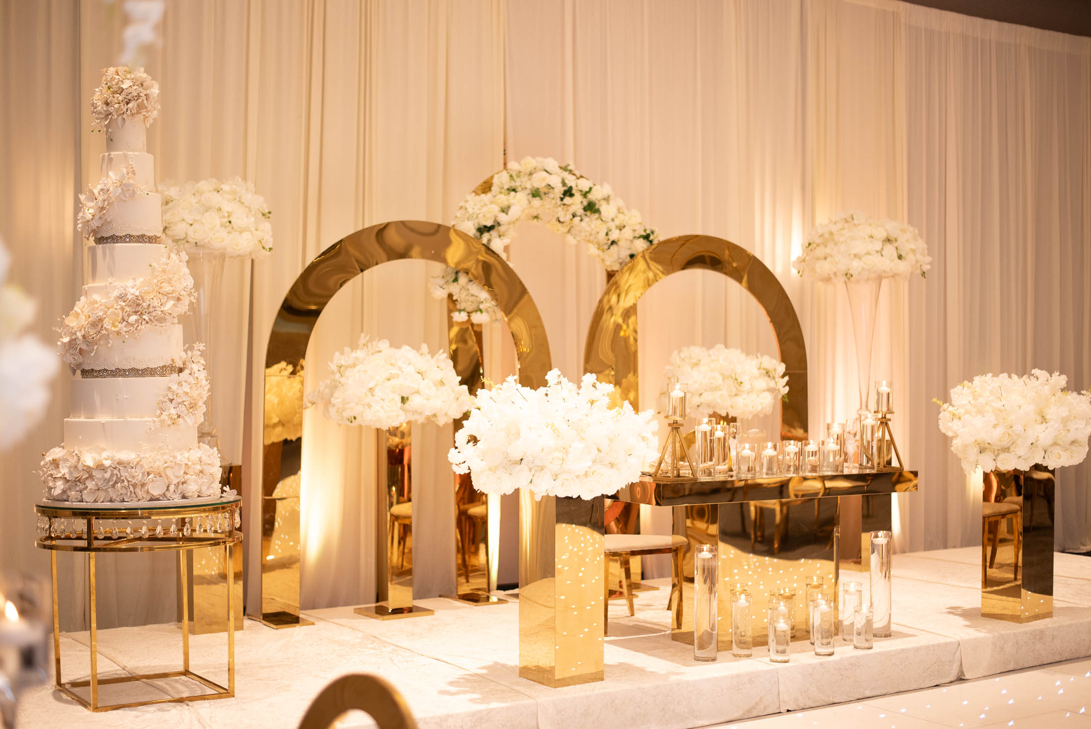 Elegant wedding setup with white flowers, gold arches, and tiered cake, decorated with floral arrangements and candles.