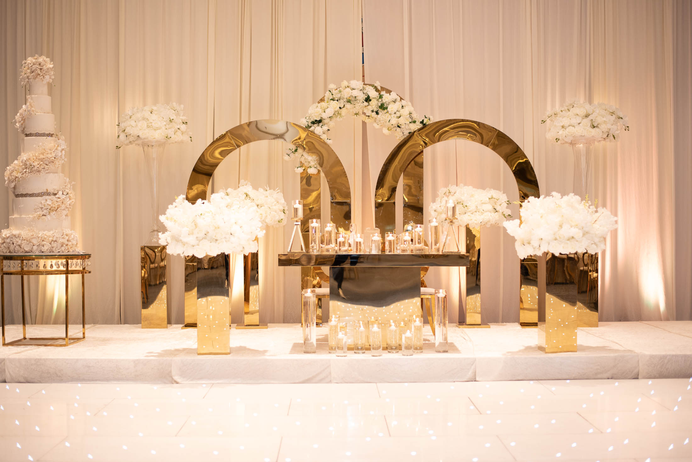Elegant wedding stage with gold and white decorations, floral arrangements, and draped curtains, set for a celebration.