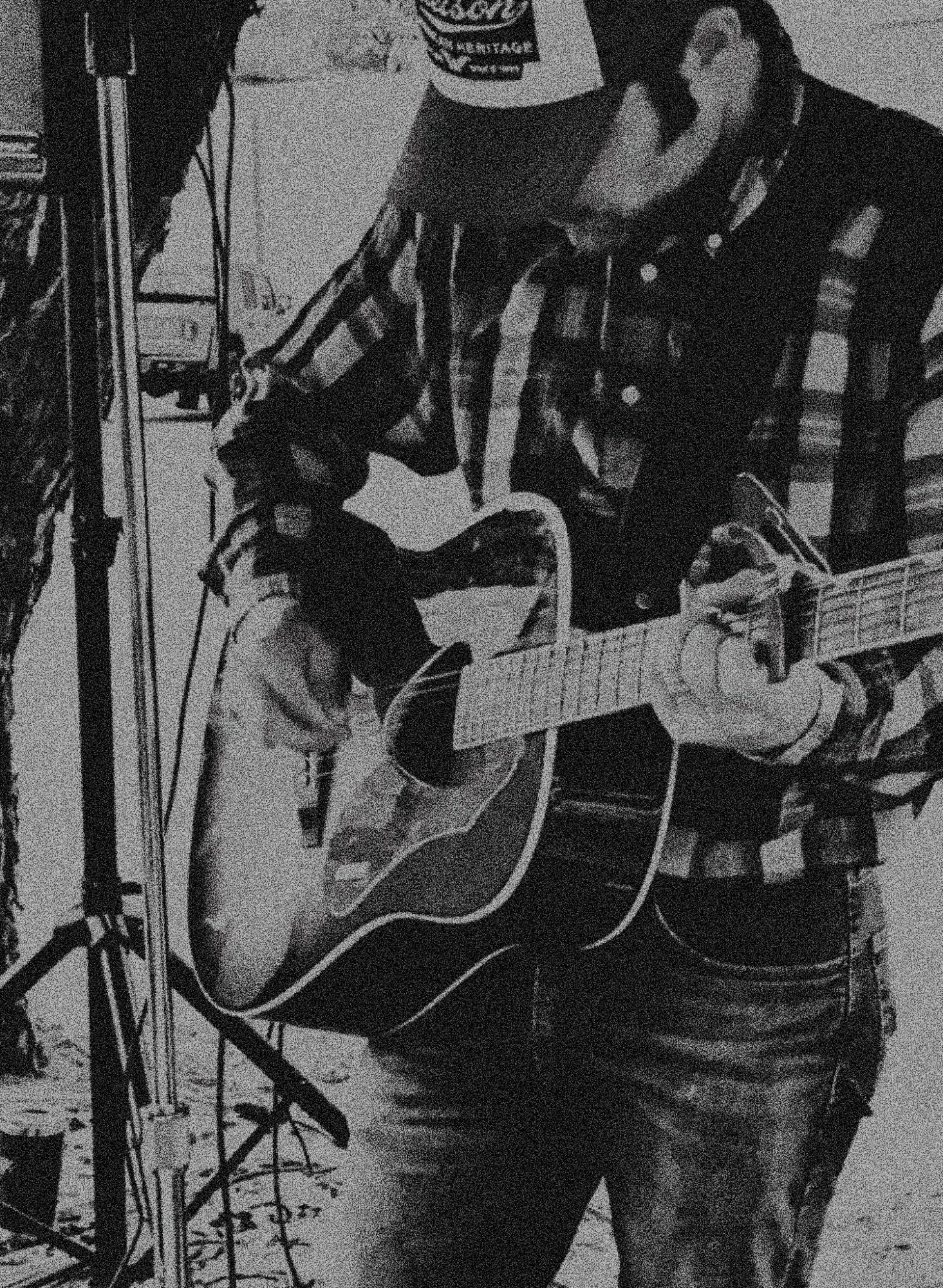 Person wearing a cap and plaid shirt playing an acoustic guitar, with equipment around them.