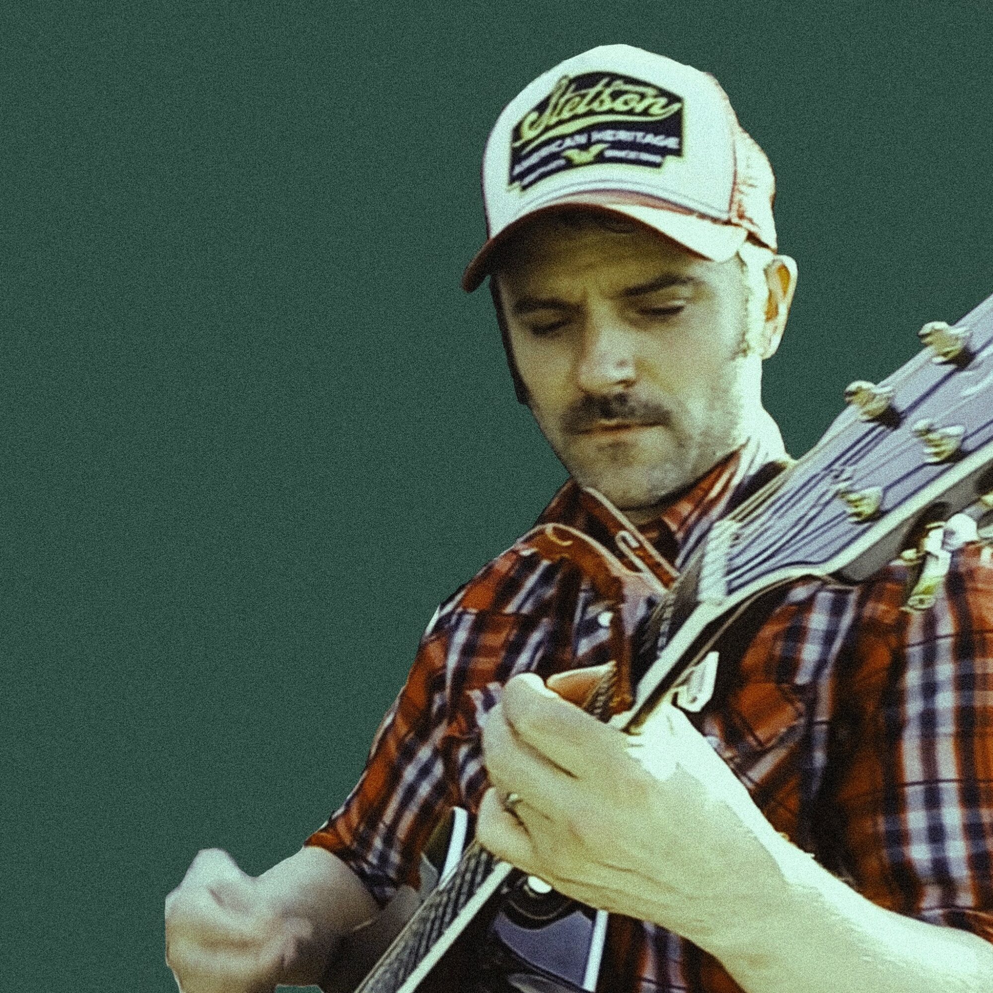 Person wearing a cap and plaid shirt playing an acoustic guitar, focused on the instrument.