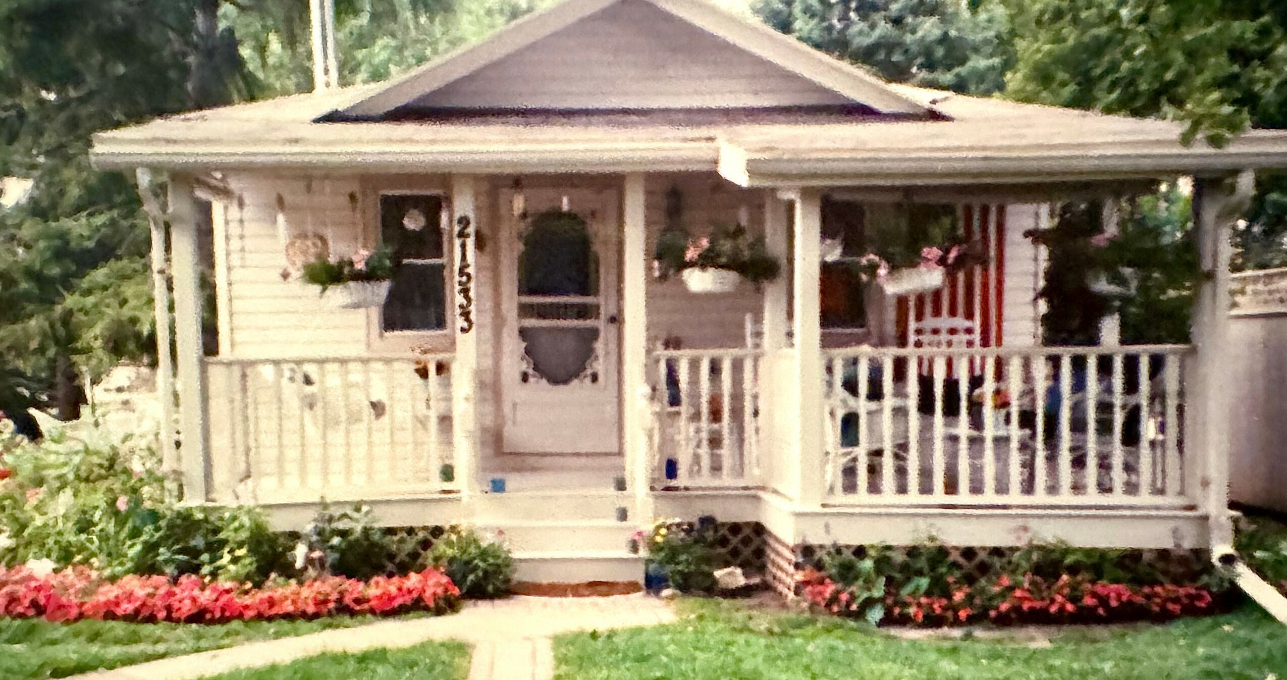 Small house with porch, white railing, hanging flower baskets, surrounded by green lawn and flower beds.
