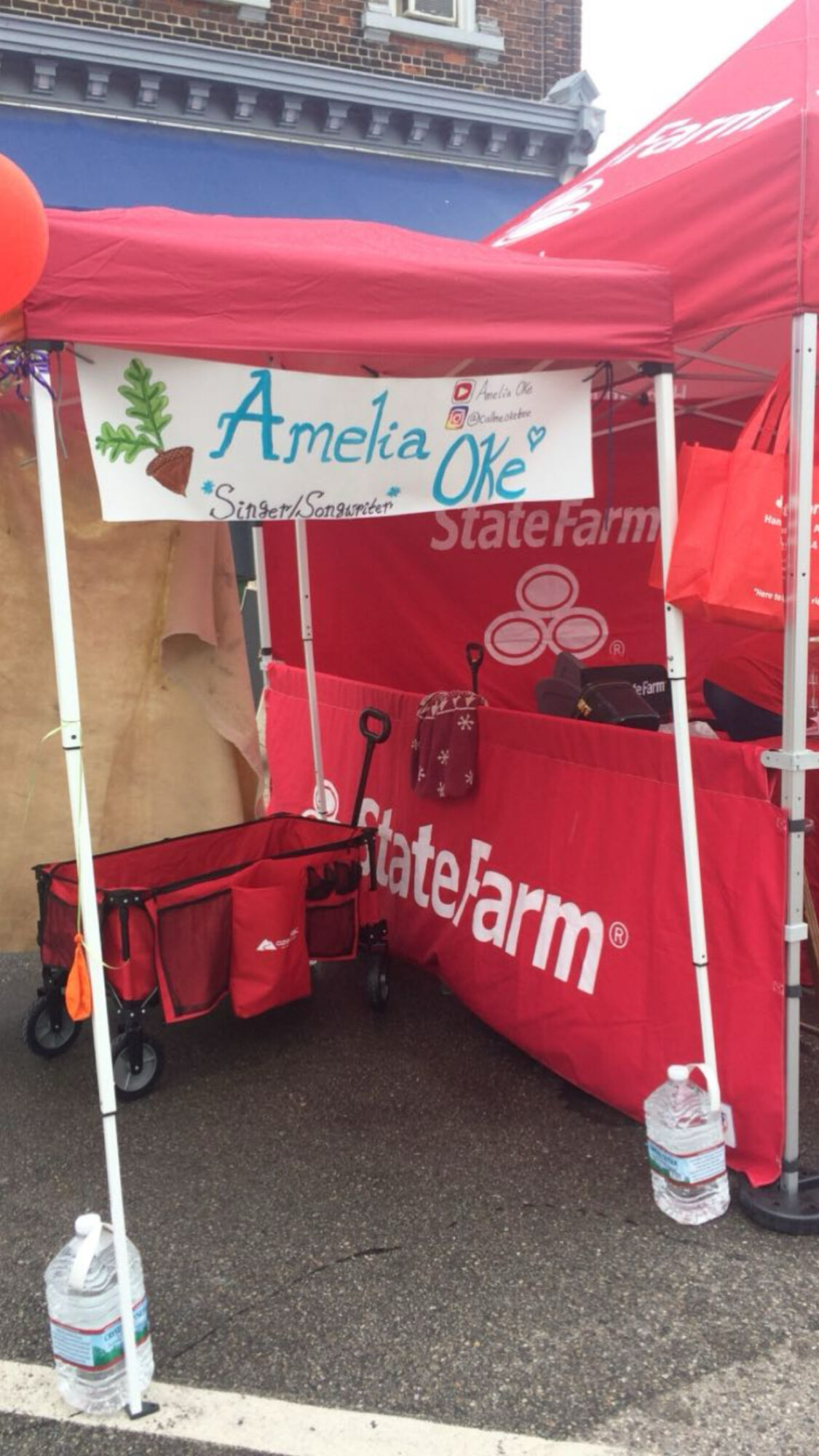 Tent with a sign reading 'Amelia Oke' and a red cooler, set up outdoors with water bottles nearby.