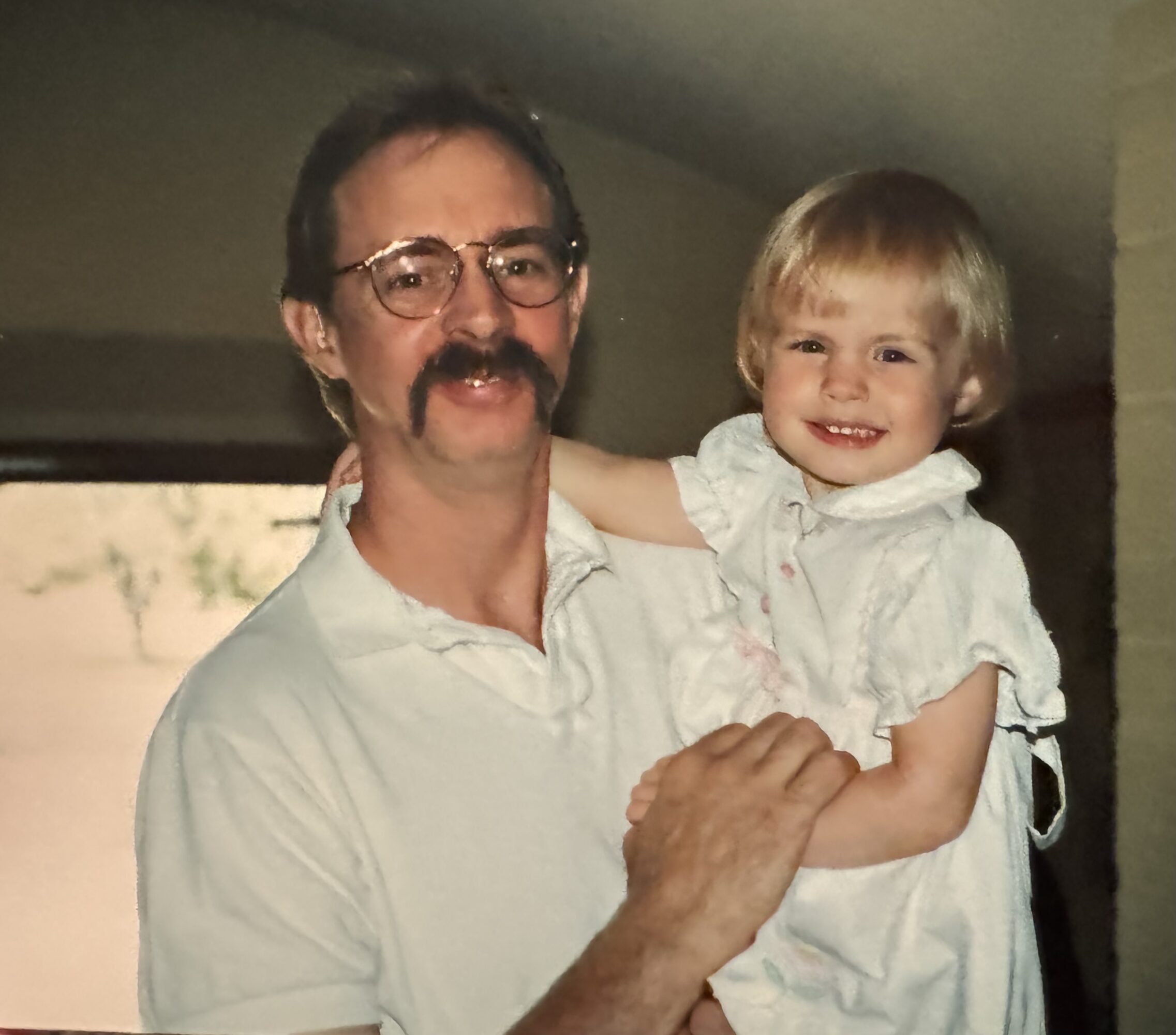 Man with glasses and a mustache holding a smiling young girl indoors.