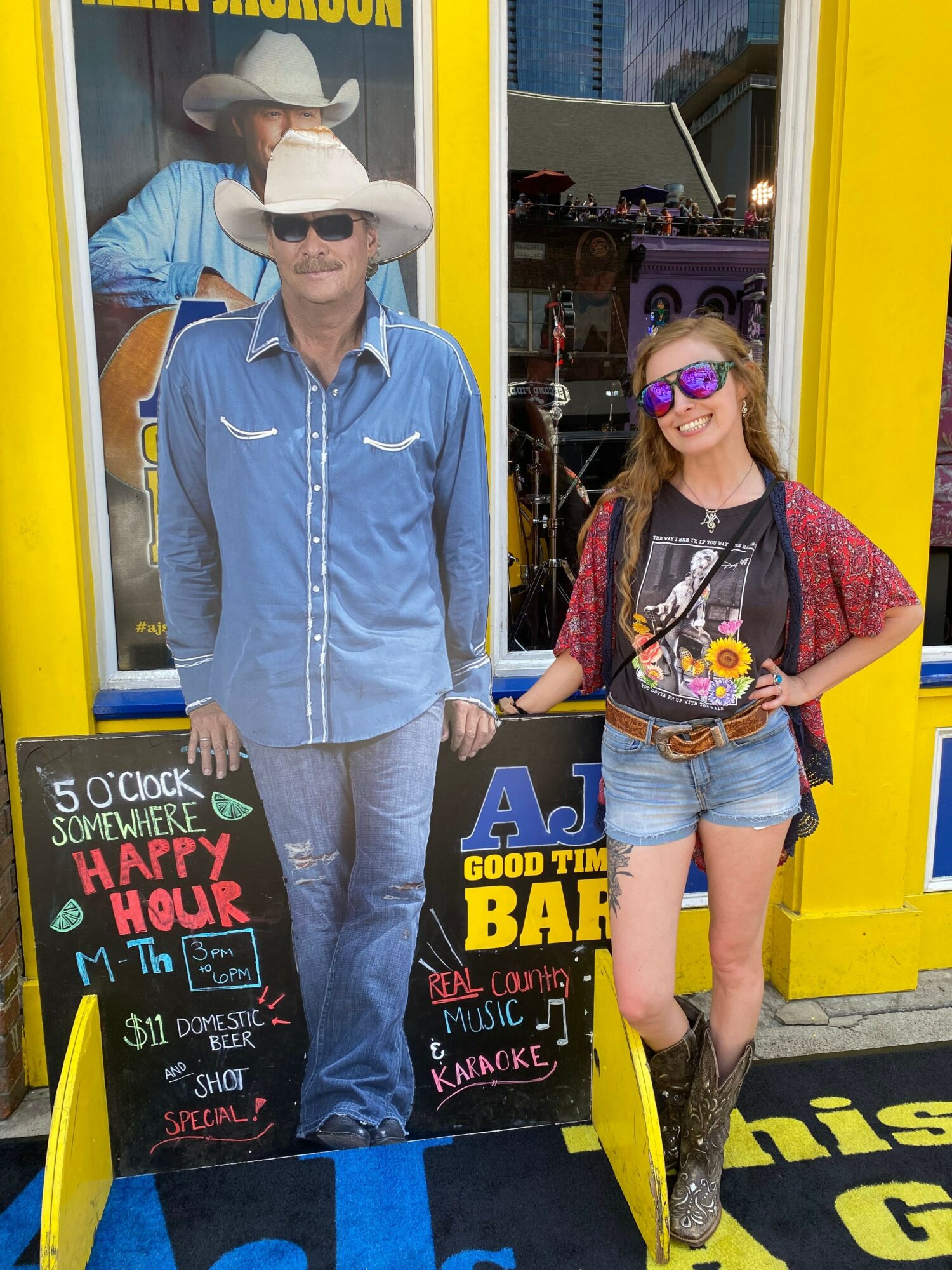 Person with purple hair and sunglasses standing next to a person in a blue hoodie and sunglasses, both in front of a yellow building with signs.
