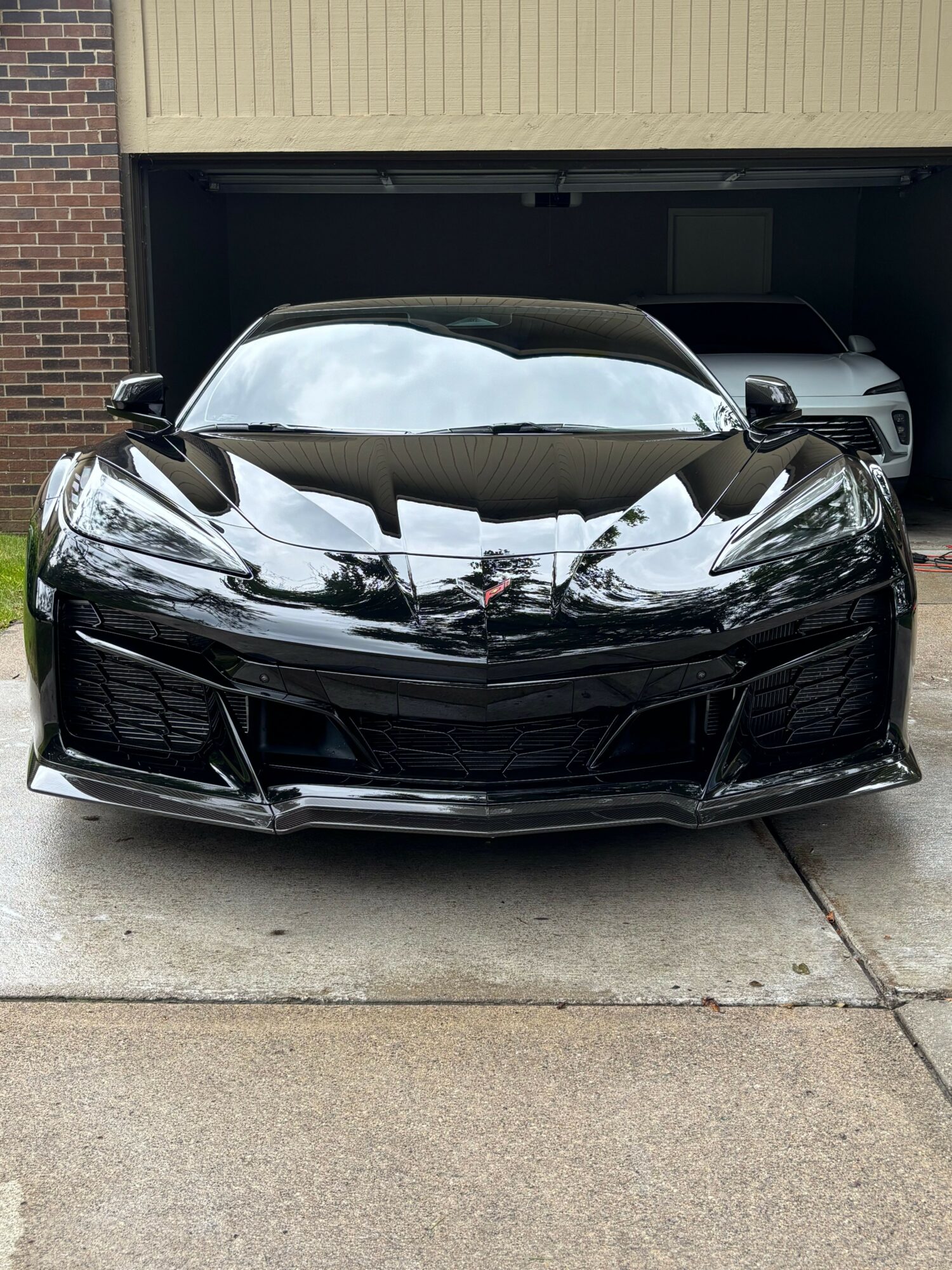 Black car parked on driveway in front of garage, with building and other vehicles visible in reflection on the car's surface.
