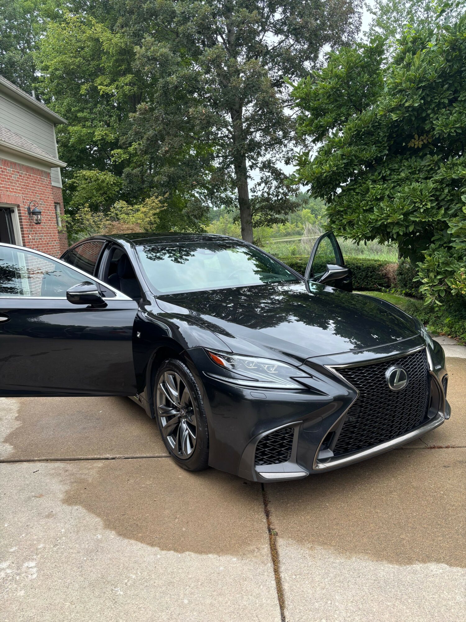 Black sports car parked on a driveway with trees and a brick house in the background.