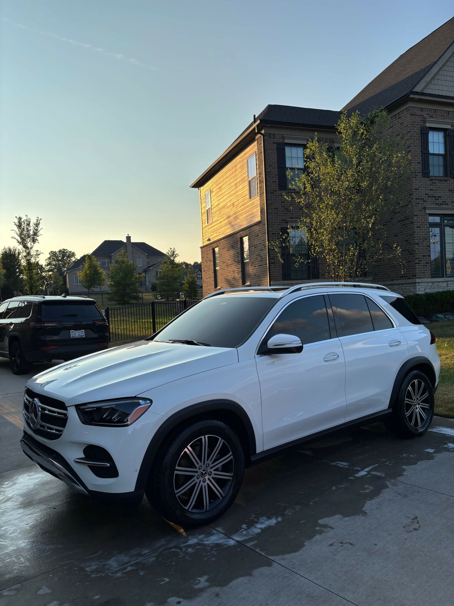 White car parked on driveway with house and trees in background, viewed sideways.