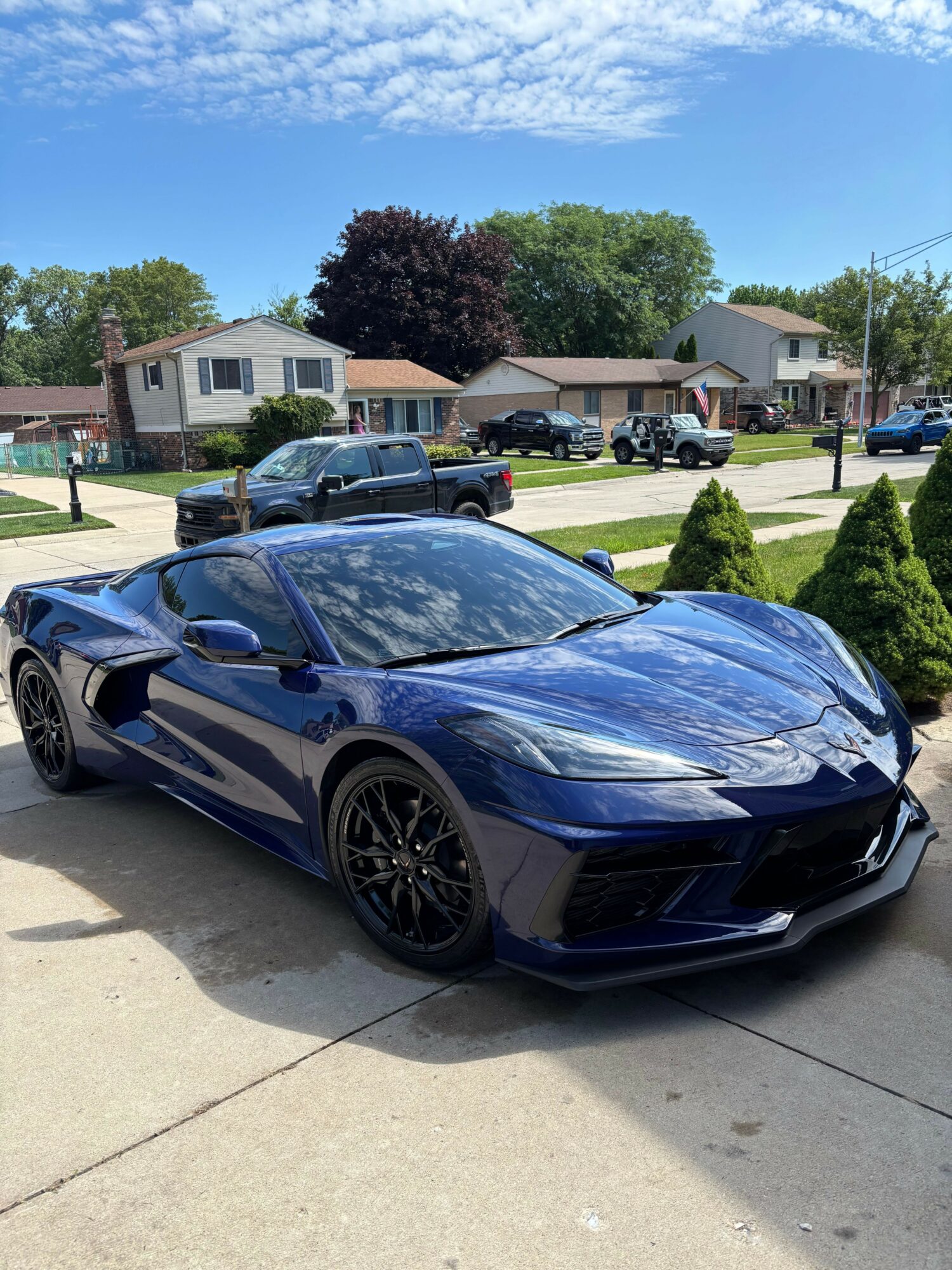 Dark blue sports car parked on a sidewalk in a residential neighborhood with trees and houses, sunny day.