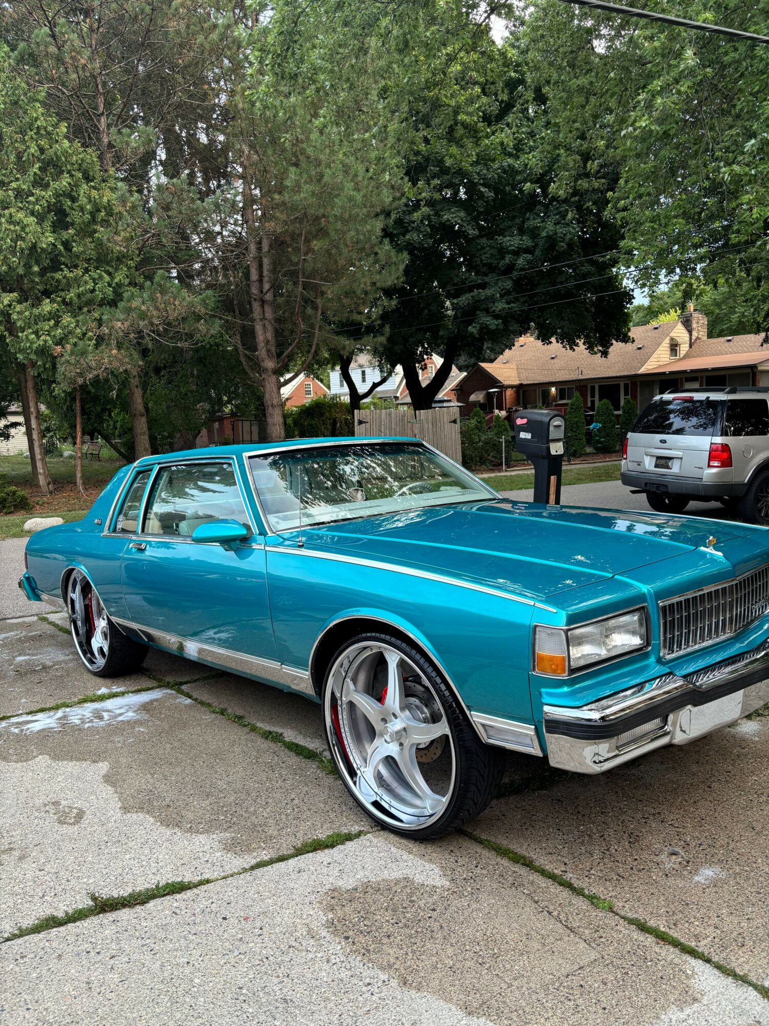 Blue vintage car parked on a sidewalk with trees and houses in the background.