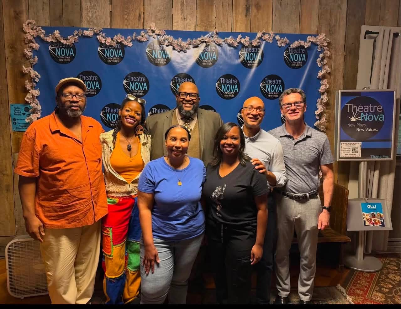 Seven people standing together in front of a blue backdrop with theater Nova logos, smiling for the photo.