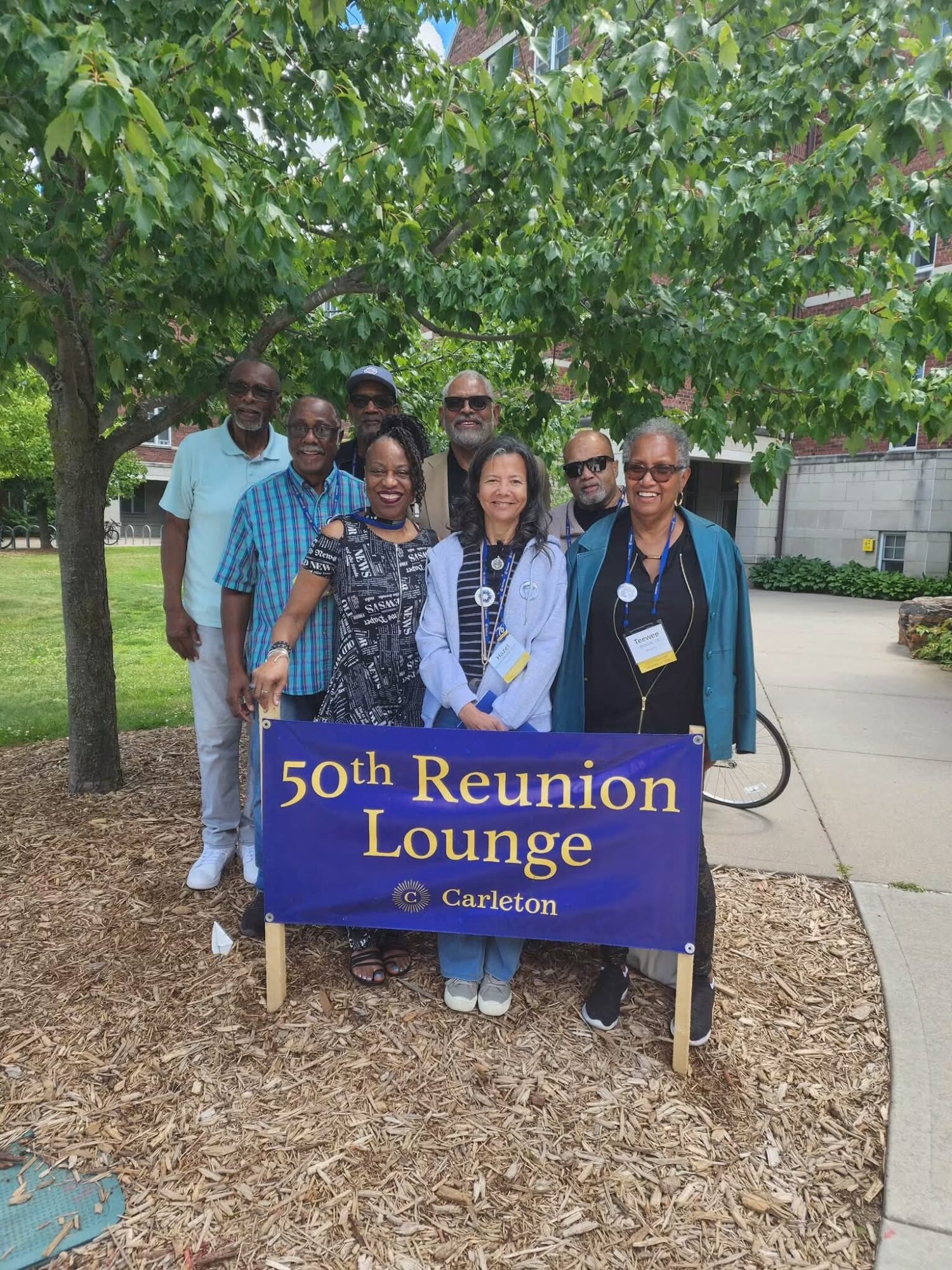 Group of eight people holding a blue banner that reads '50th Reunion Lounge' in a park with trees and a sidewalk