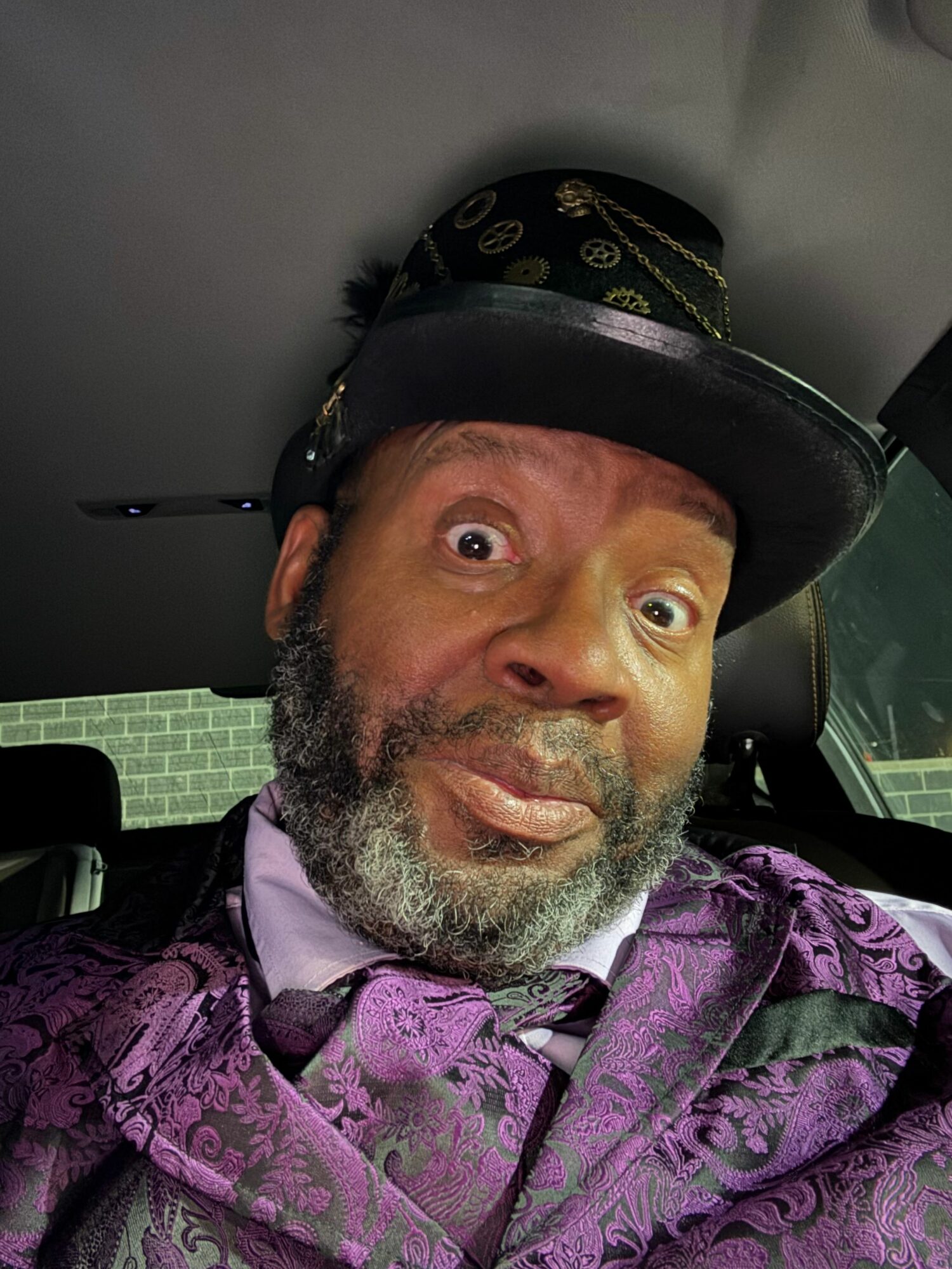 Man with a beard and mustache wearing a black hat and purple patterned jacket, sitting in a vehicle.