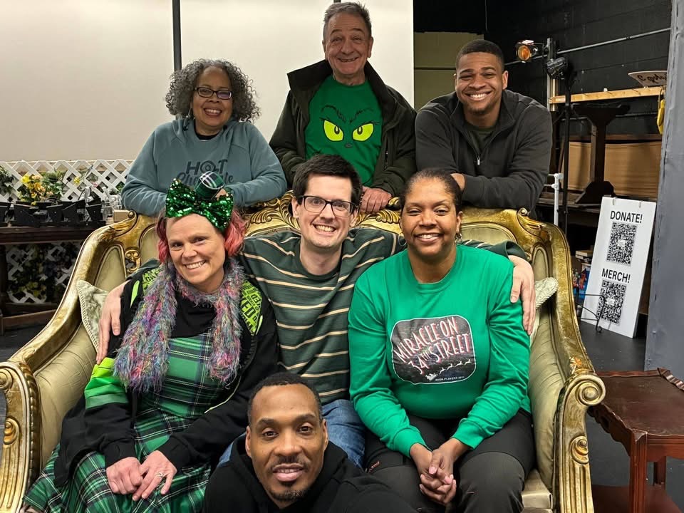 Six people smiling and sitting on a vintage sofa, with three standing behind them, in an indoor setting.