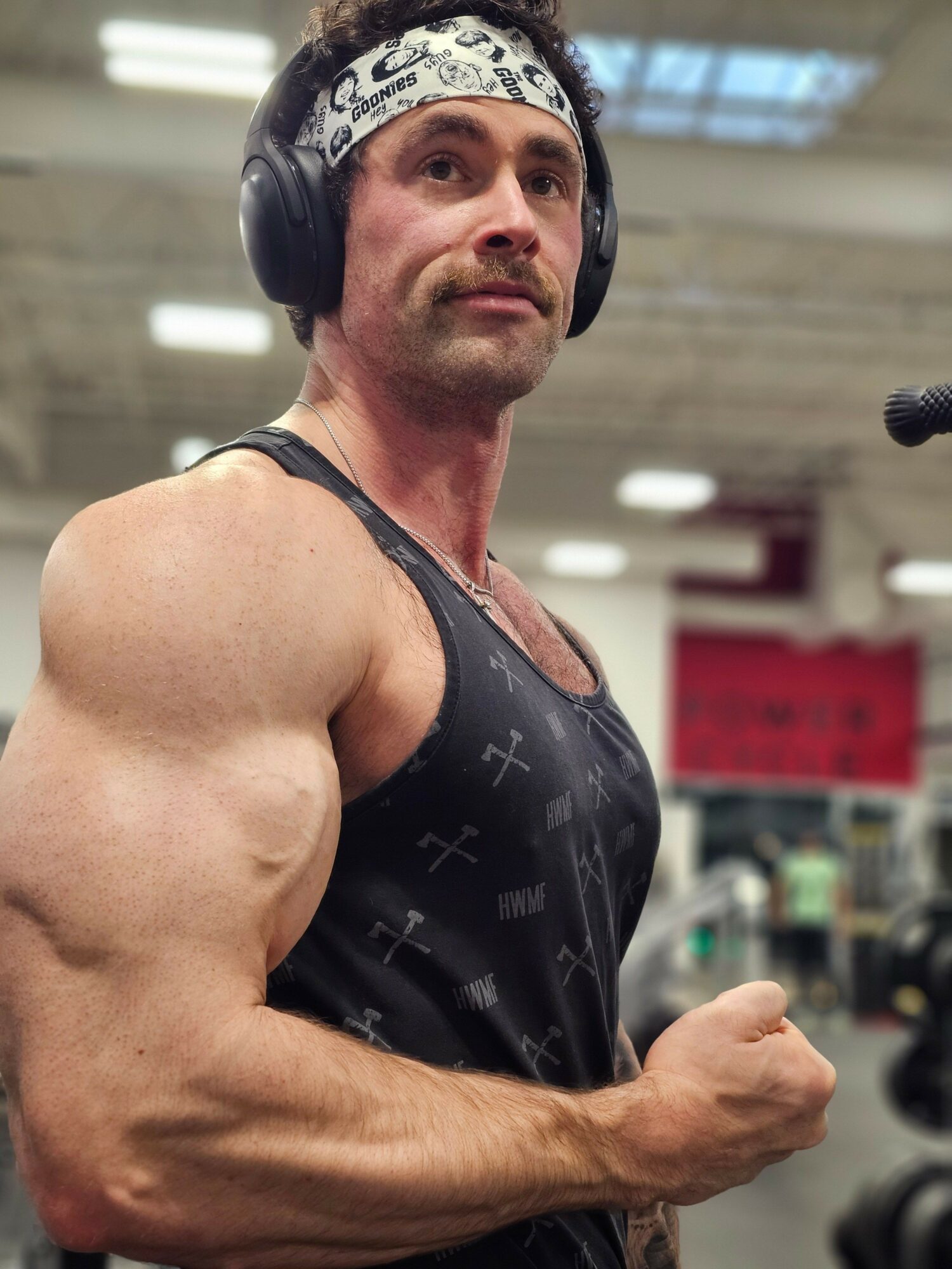 A muscular man wearing a black tank top and a bandana, flexing his arm in a gym.