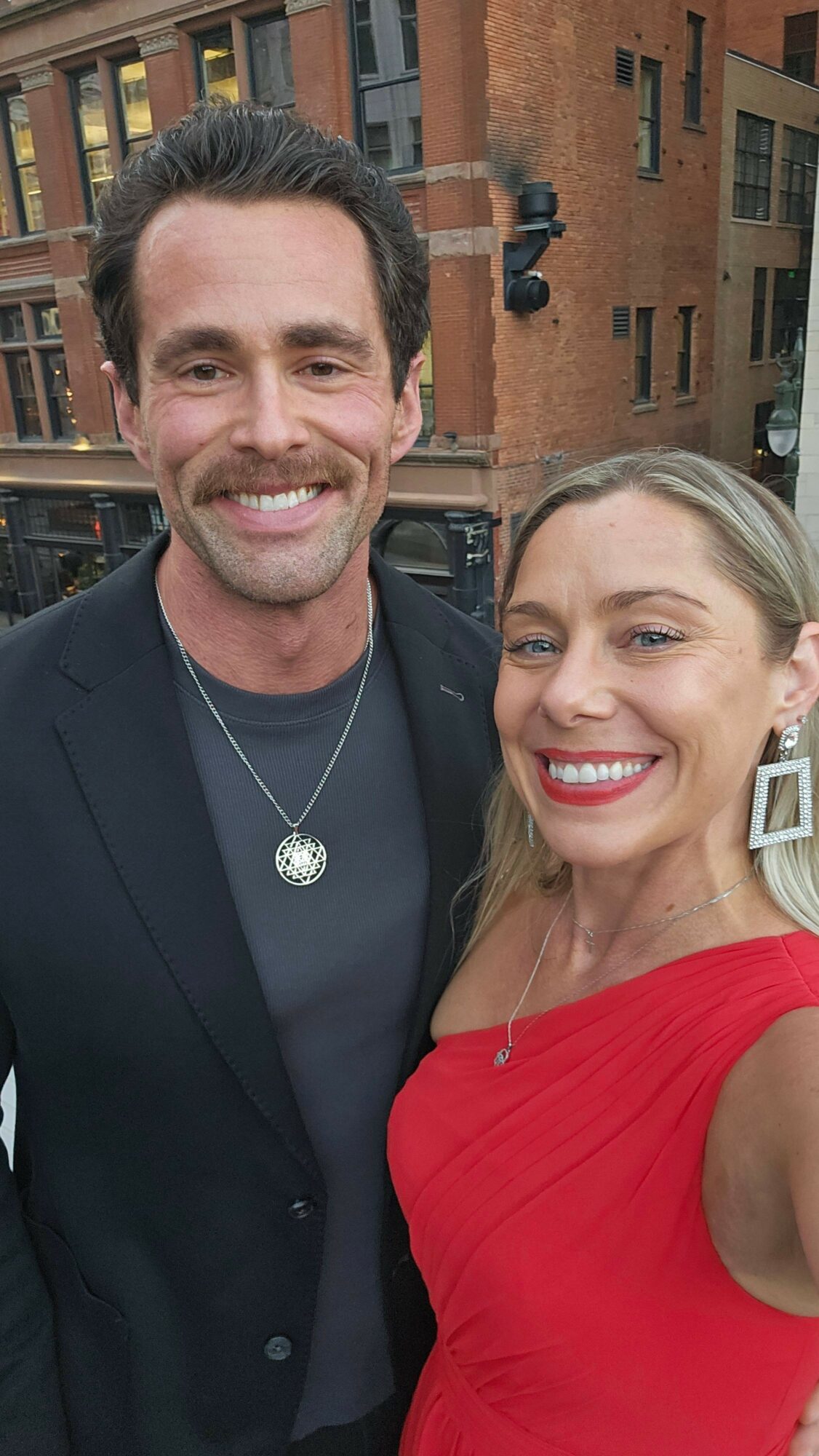 Two smiling people, a man in a dark suit and a woman in a red dress, standing outdoors in front of brick buildings.