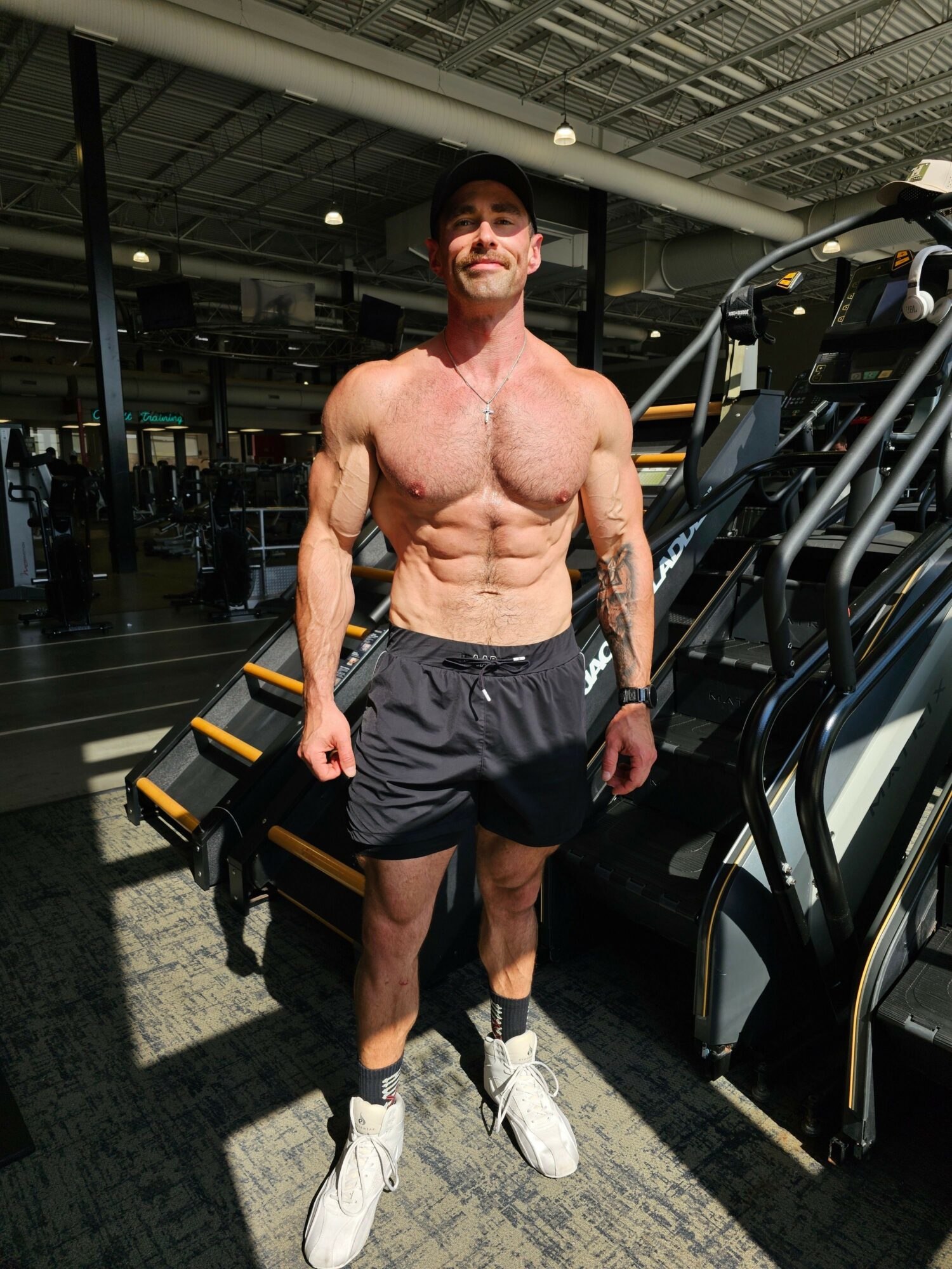 A muscular man with defined abs and arms in a gym, wearing black shorts and white shoes, standing near gym equipment.
