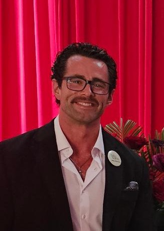 Man with glasses, dark hair, and a mustache, smiling, wearing a black blazer and white shirt, standing in front of red curtains.