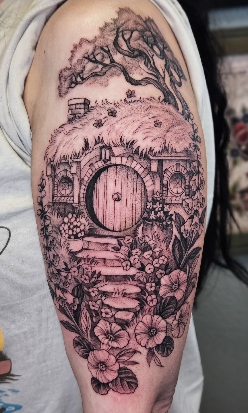 Tattoo of a house with a thatched roof, surrounded by flowers, trees, and a pathway, on a person's upper arm.