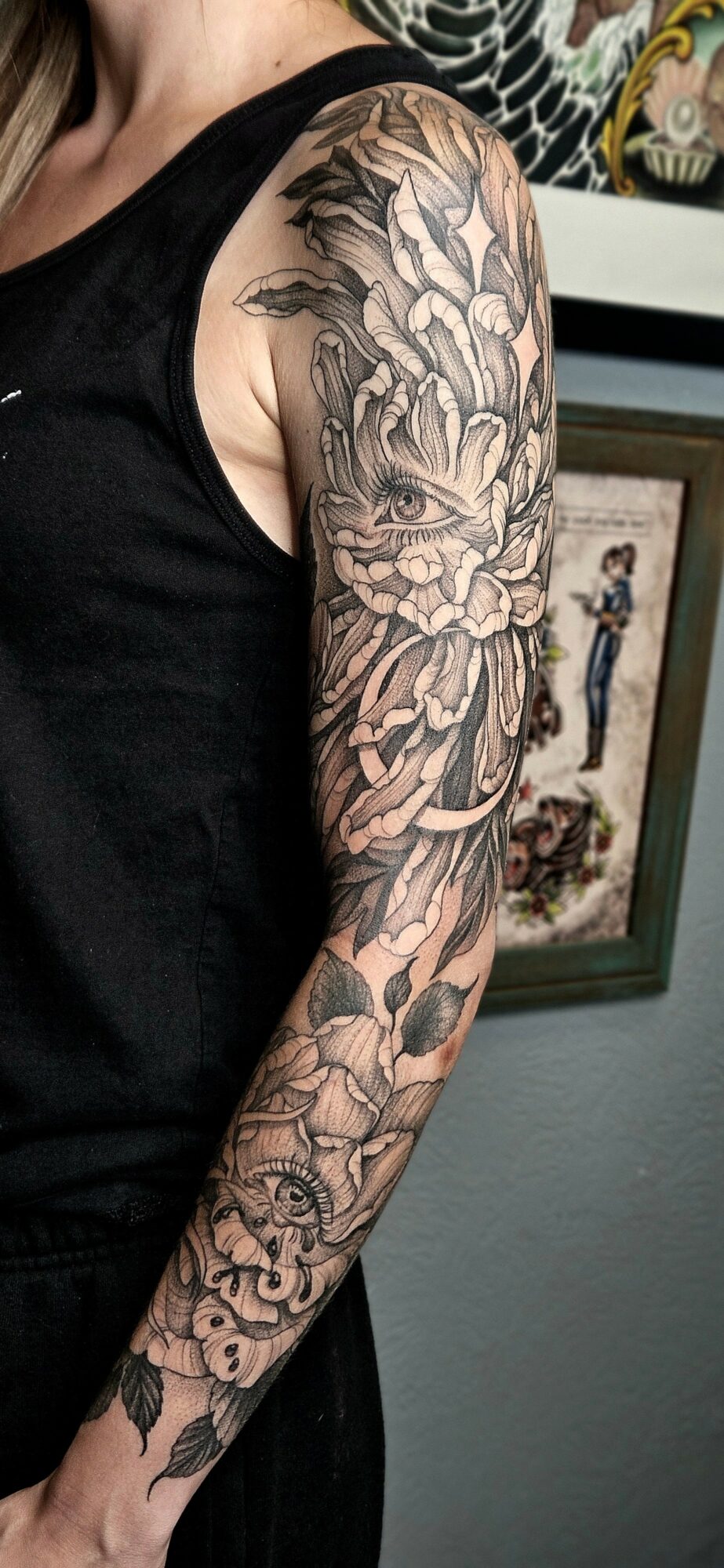 Tattooed arm with floral and animal designs, person wearing a black sleeveless shirt, background with framed artwork.
