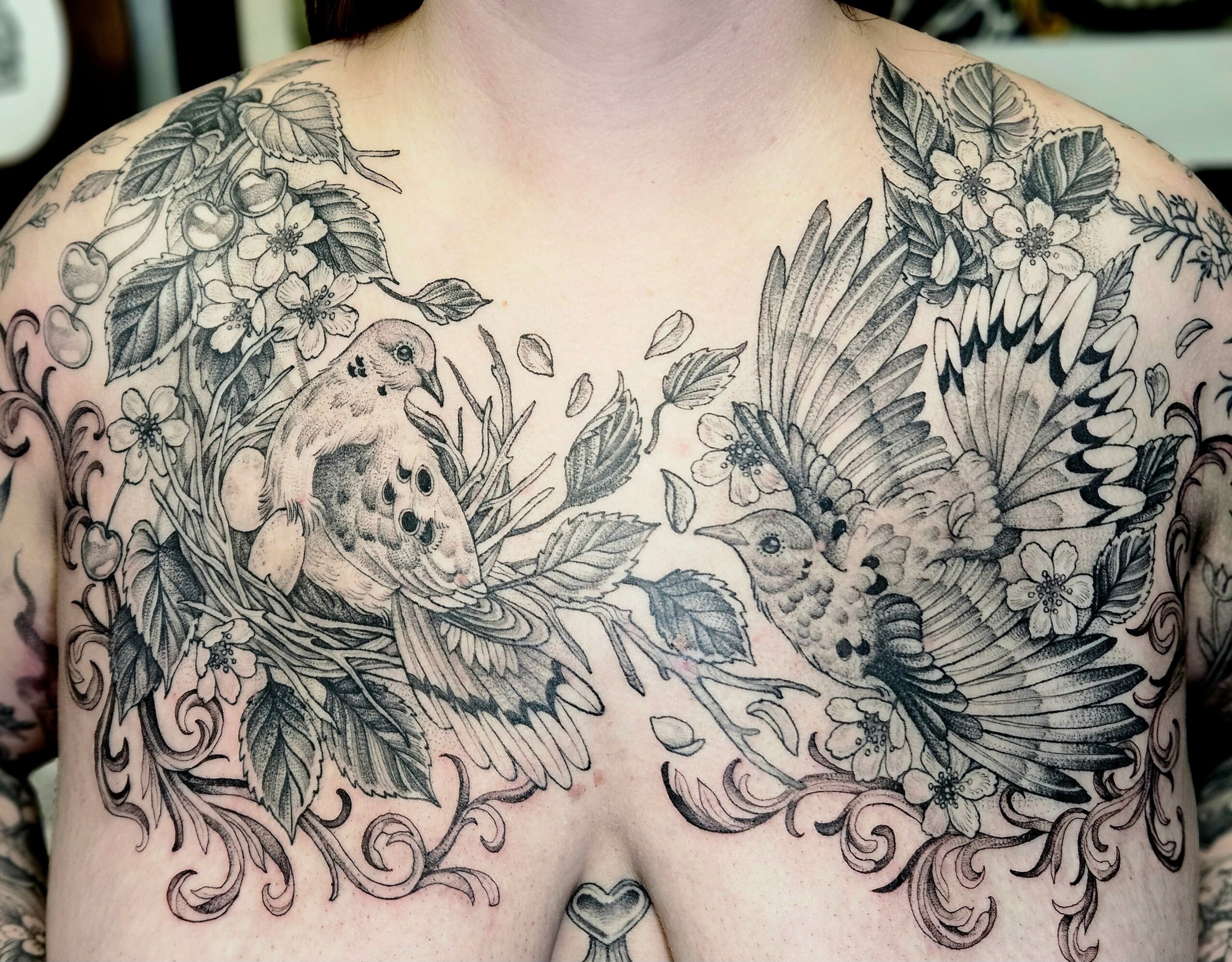 Tattooed chest with detailed birds, flowers, and leaves in black ink, covering shoulders and upper chest area.