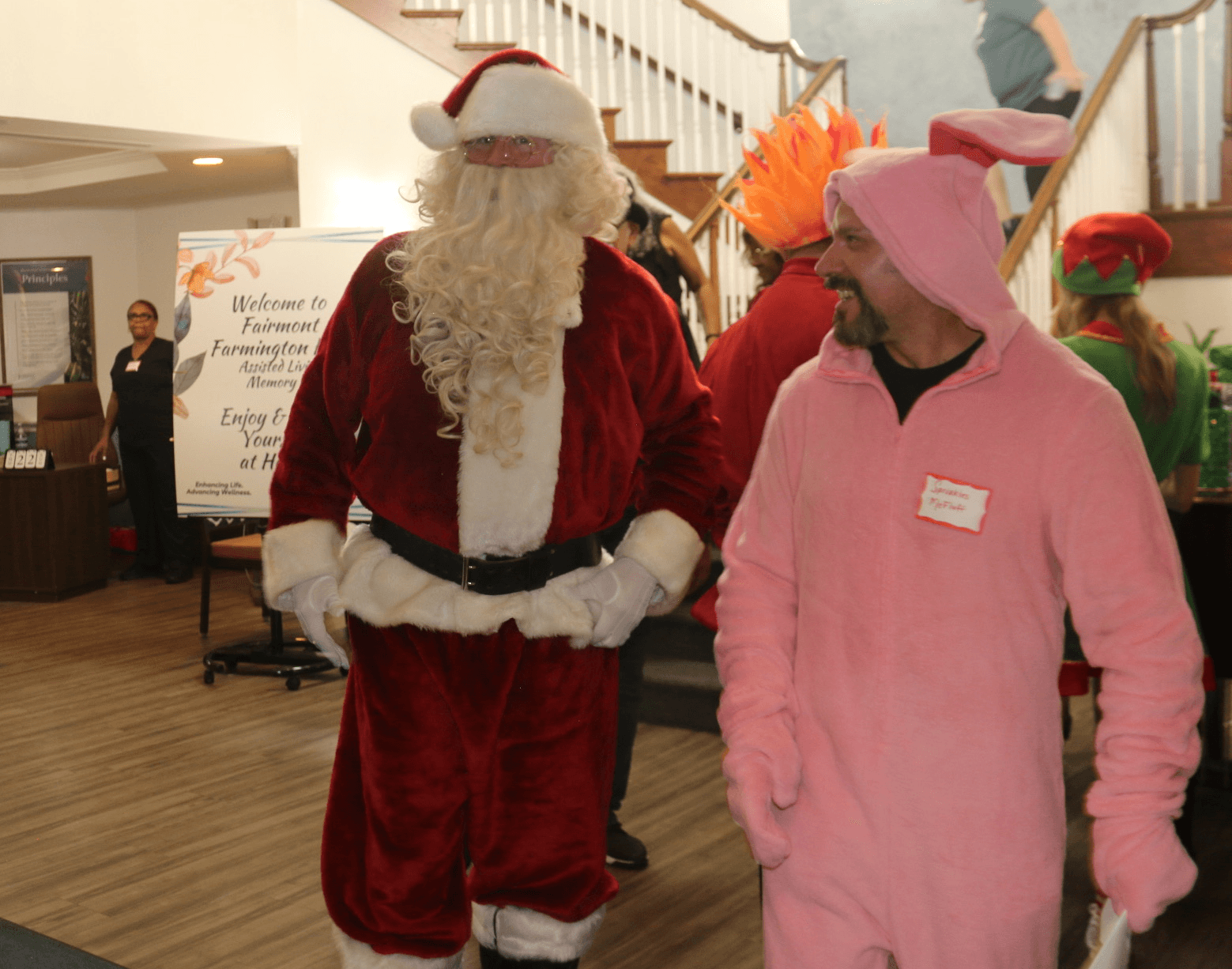 Santa Claus in a red suit and hat walking with a person in a pink costume, indoors with a staircase in background.