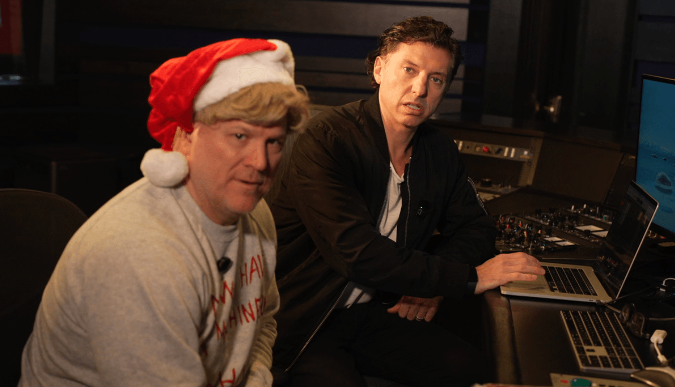 Two men sitting at a desk with audio equipment, one wearing a Santa hat, in a recording studio.