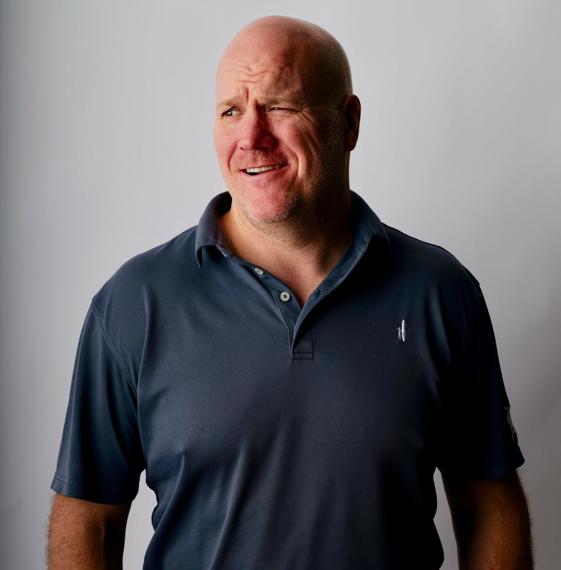 Bald man with a puzzled expression, wearing a dark polo shirt, standing against a plain light background.