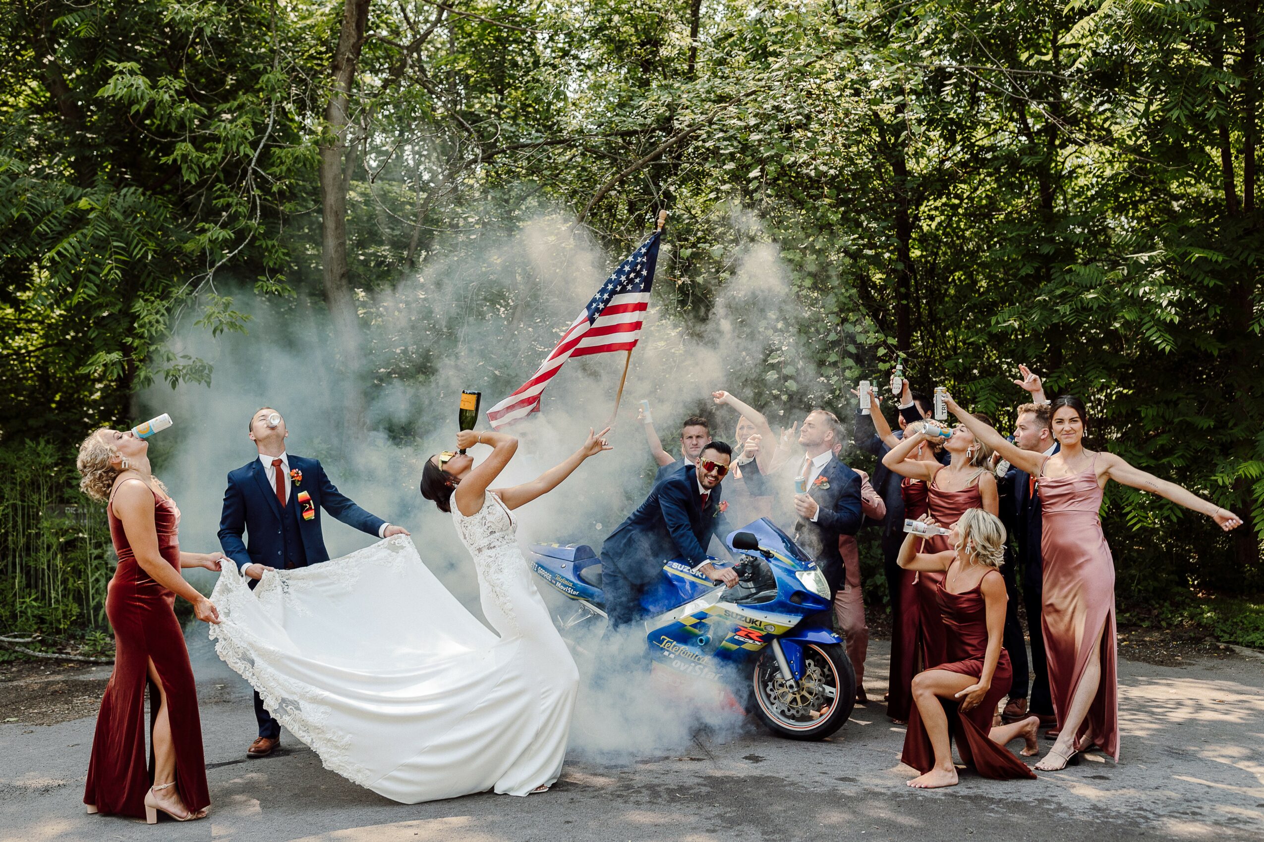 People celebrating outdoors with a motorcycle, a person holding an American flag, and women in formal dresses, some dancing.