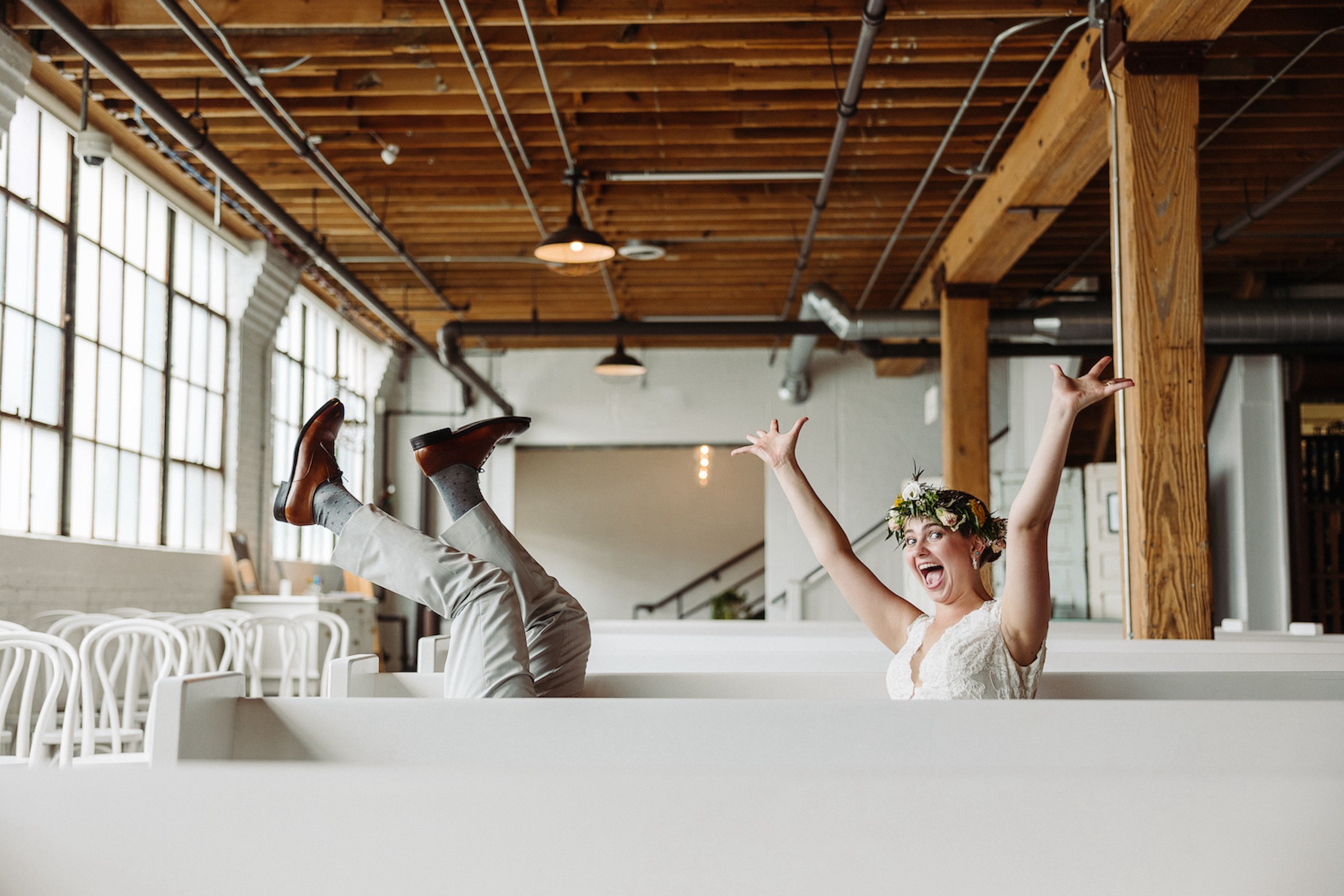 Person with arms raised, wearing glasses and a floral headband, sitting in a bright industrial-style space with wooden beams and large windows.