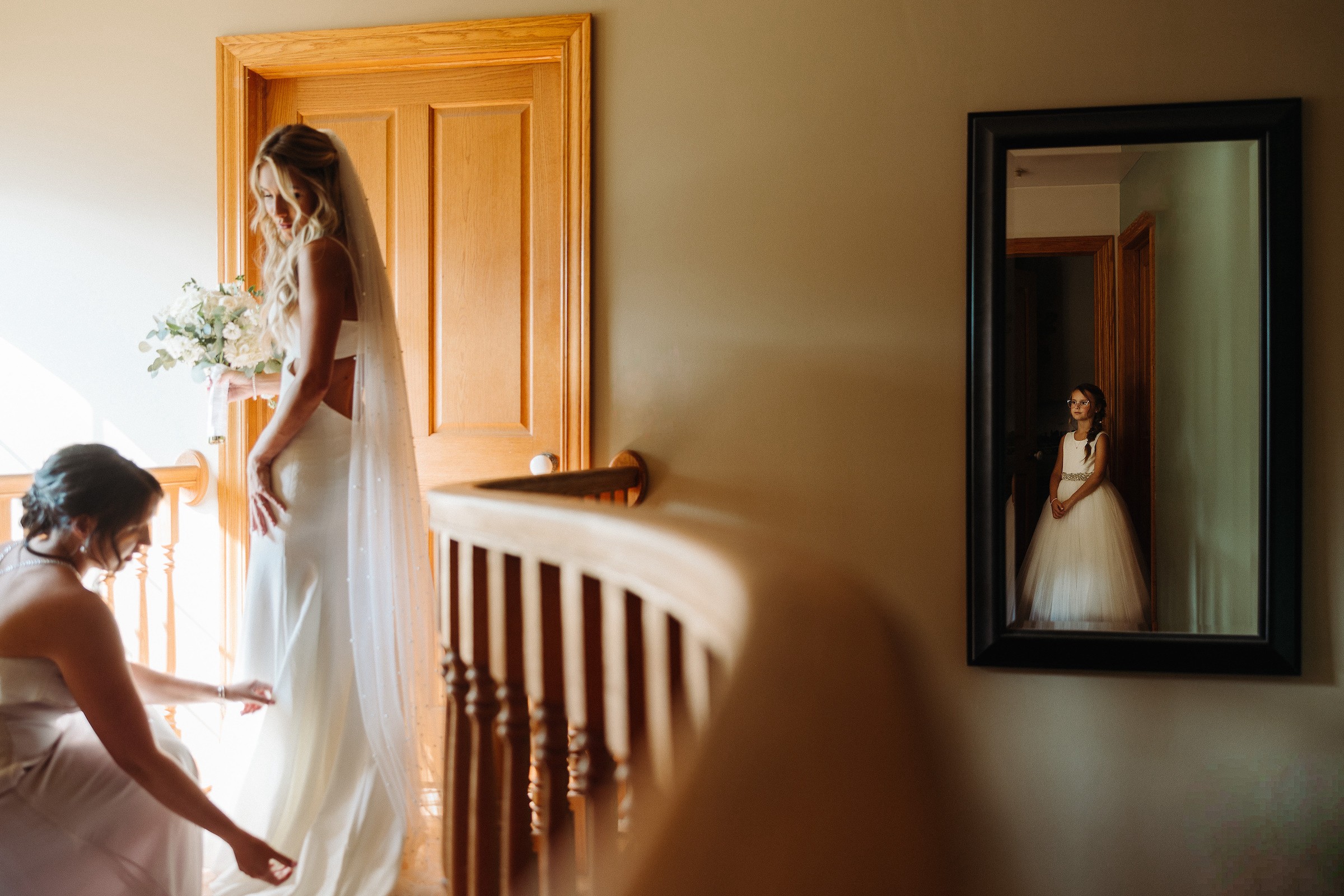 A woman in a white dress stands near a door, reflected in a mirror on the wall, with a person kneeling nearby.