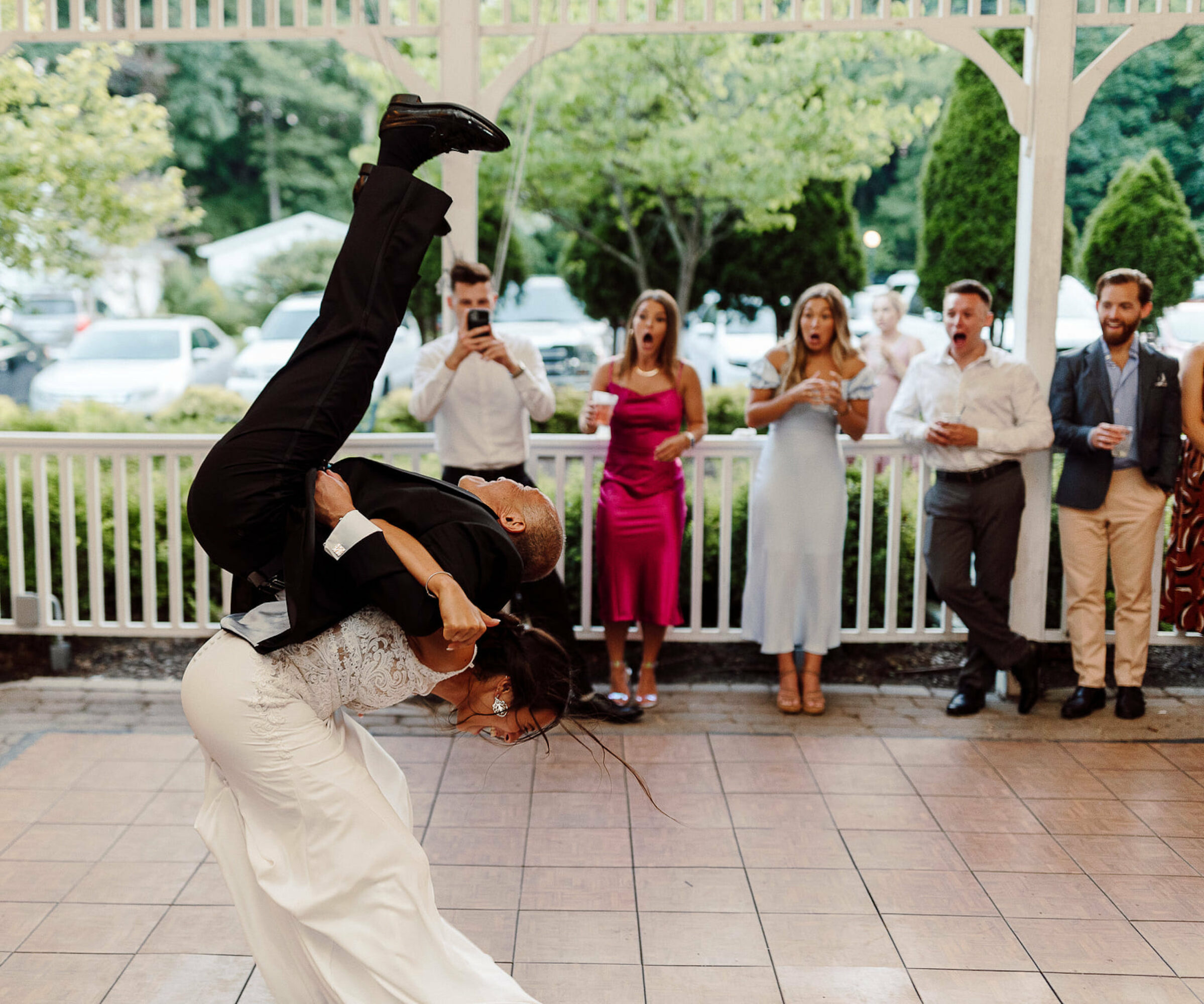 Two people dancing, one person upside down, with five onlookers clapping and taking photos outdoors on a porch.