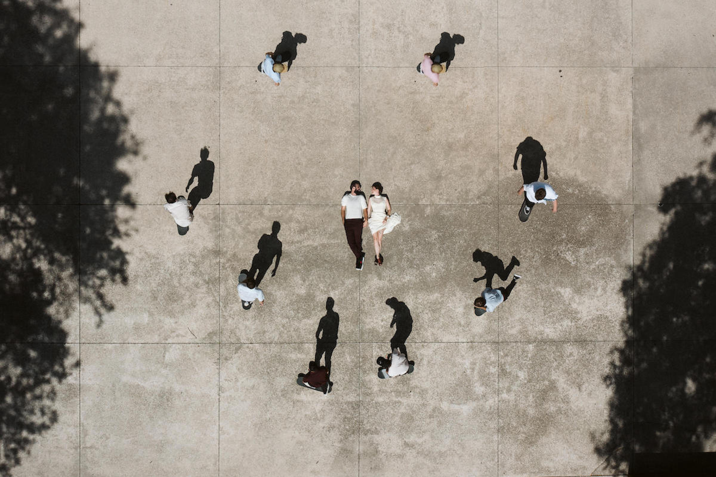 People walking on a large concrete area with trees on sides, viewed from above, some carrying bags or backpacks.
