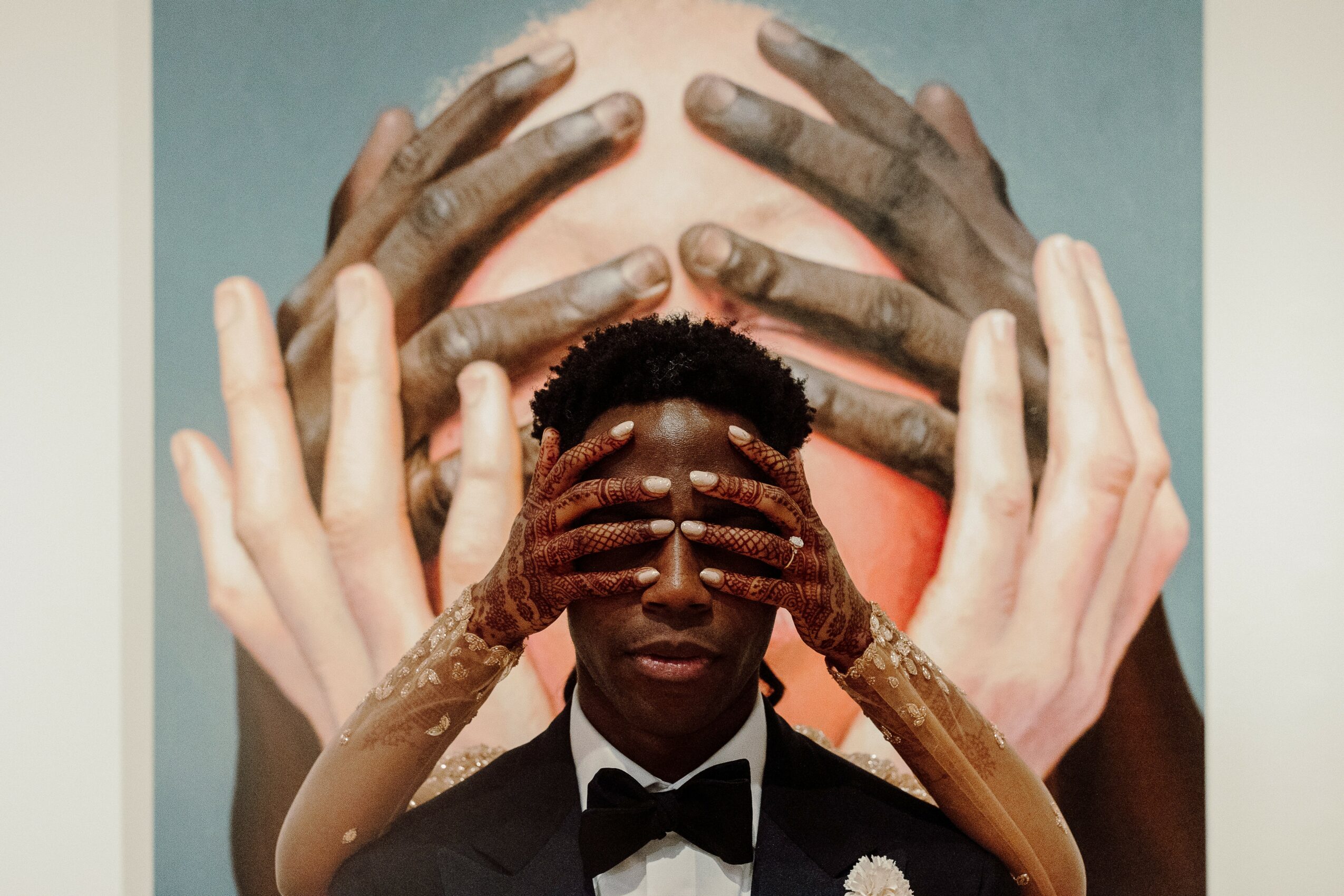 Person with dark skin and short hair, eyes covered by hands, surrounded by multiple hands reaching towards face, background with large hands.