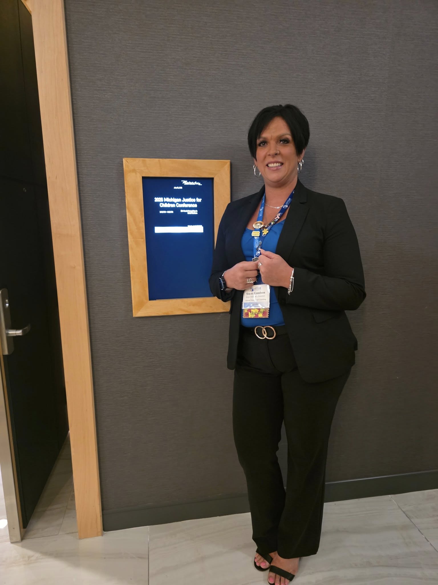 Woman in black suit standing next to a digital display on a gray wall, smiling, holding a lanyard, in an indoor setting.