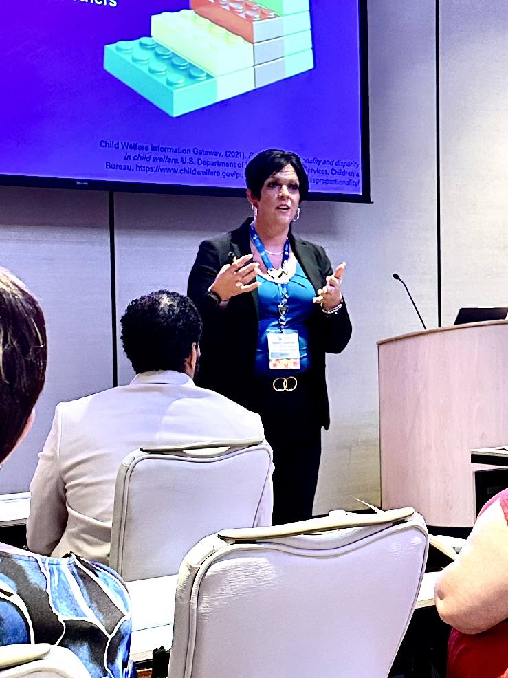 Woman giving a presentation in a conference room with a screen behind her, audience seated facing her.