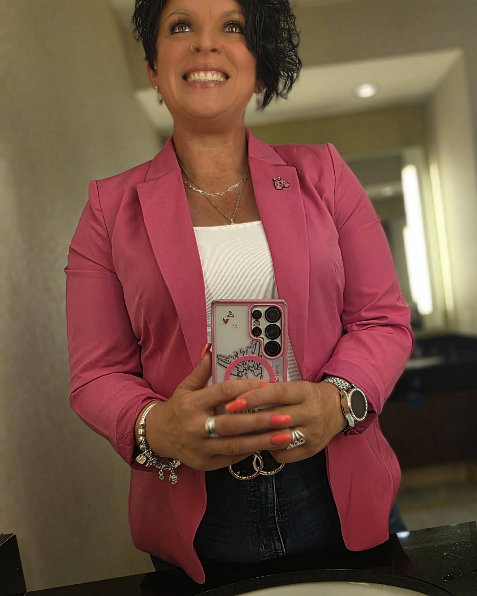 Woman in pink blazer taking a mirror selfie, smiling, in a room with a doorway in the background.