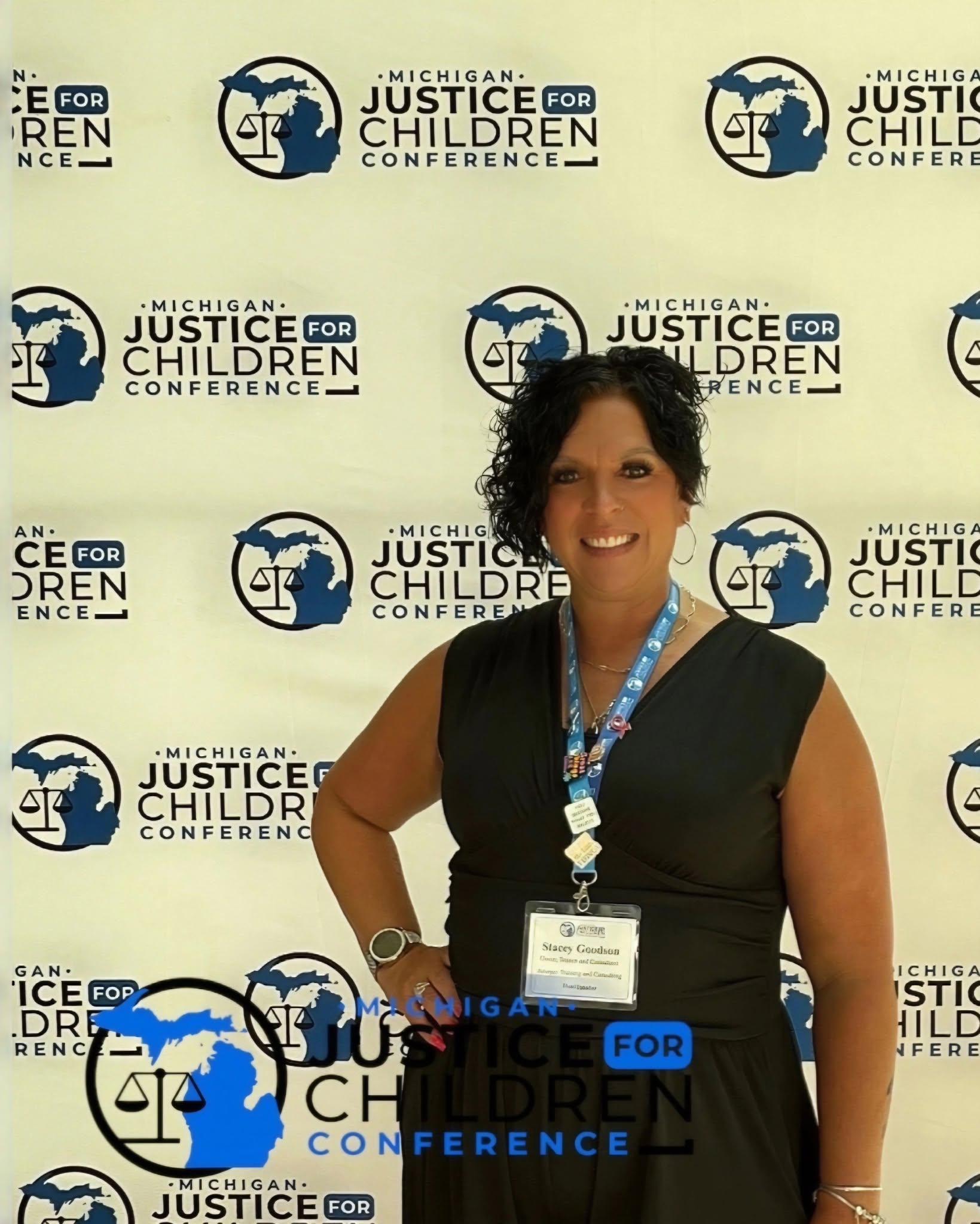 Woman smiling in front of Michigan Justice for Children Conference backdrop, wearing a black dress and conference badge.