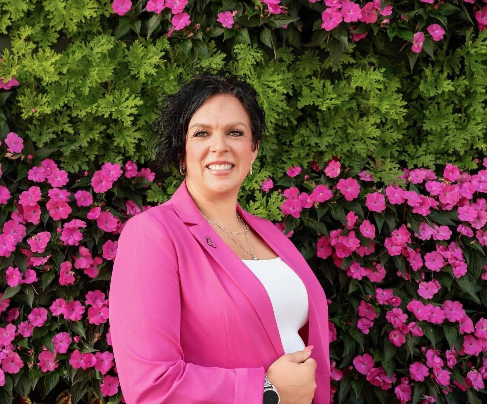 Woman with short dark hair smiling in front of pink and purple flowers and green foliage.