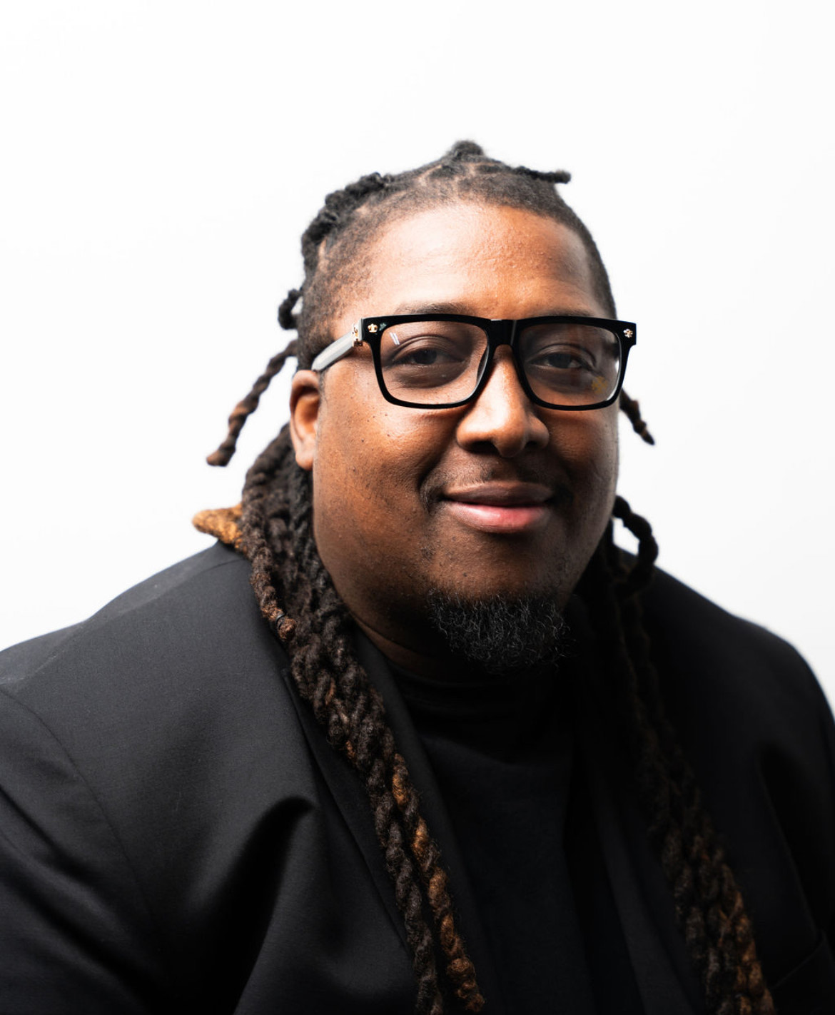 Person with glasses and dreadlocks smiling, wearing a black jacket, against a white background.