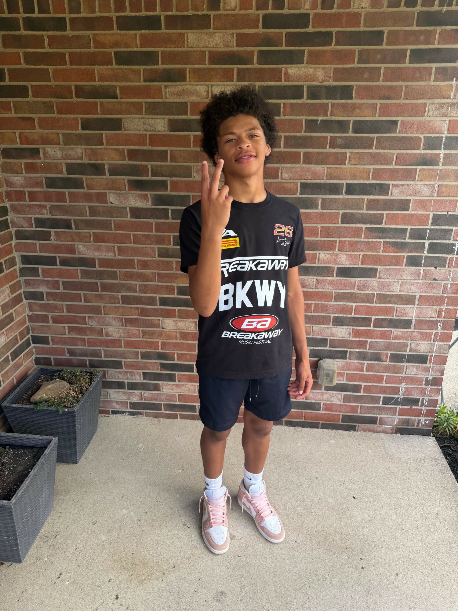 Young woman with curly hair standing outdoors against a brick wall, talking on a cellphone, wearing a black sports shirt, shorts, and sneakers.