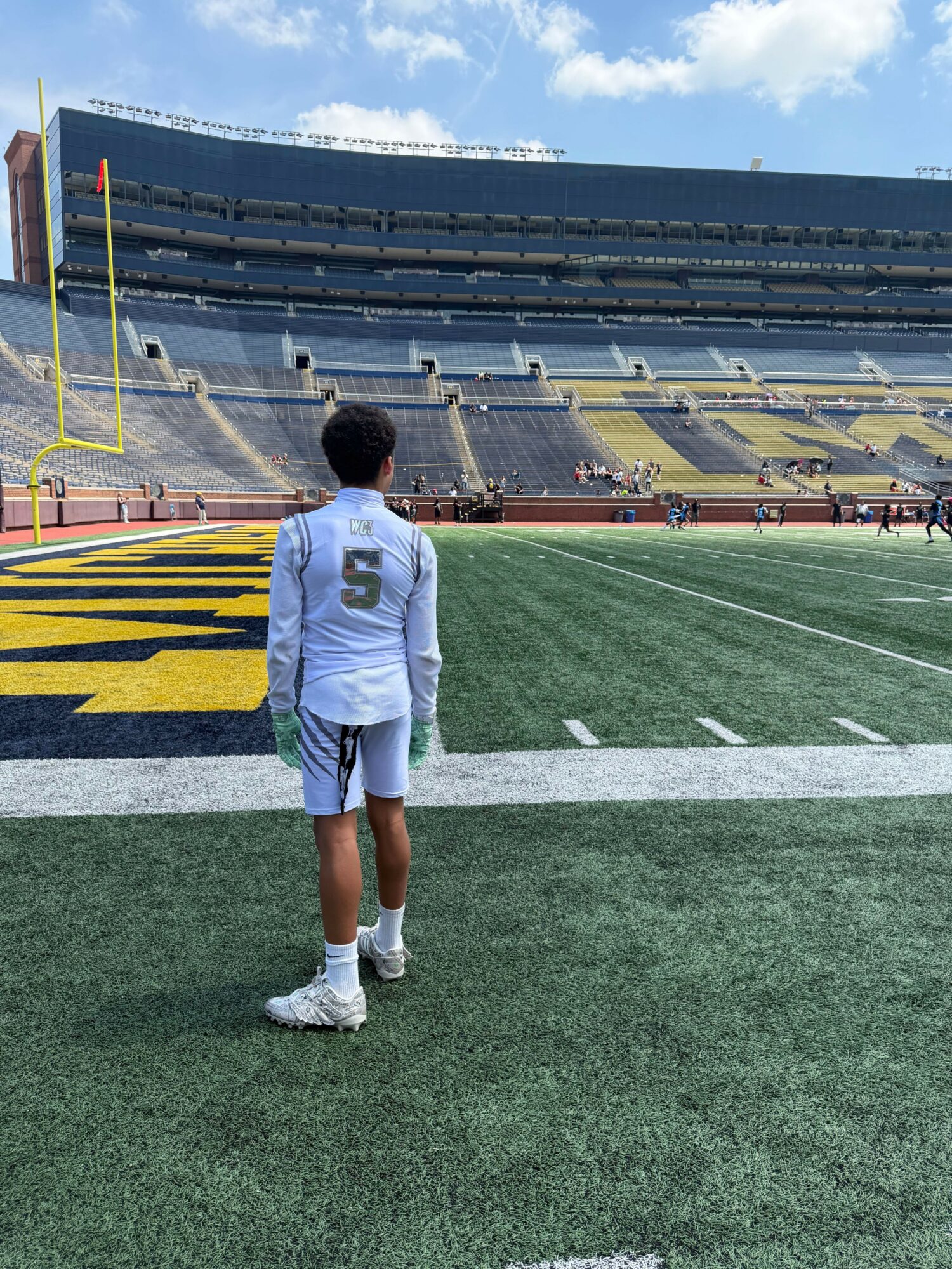 Person in white sports uniform standing on a football field with stadium seats in the background.