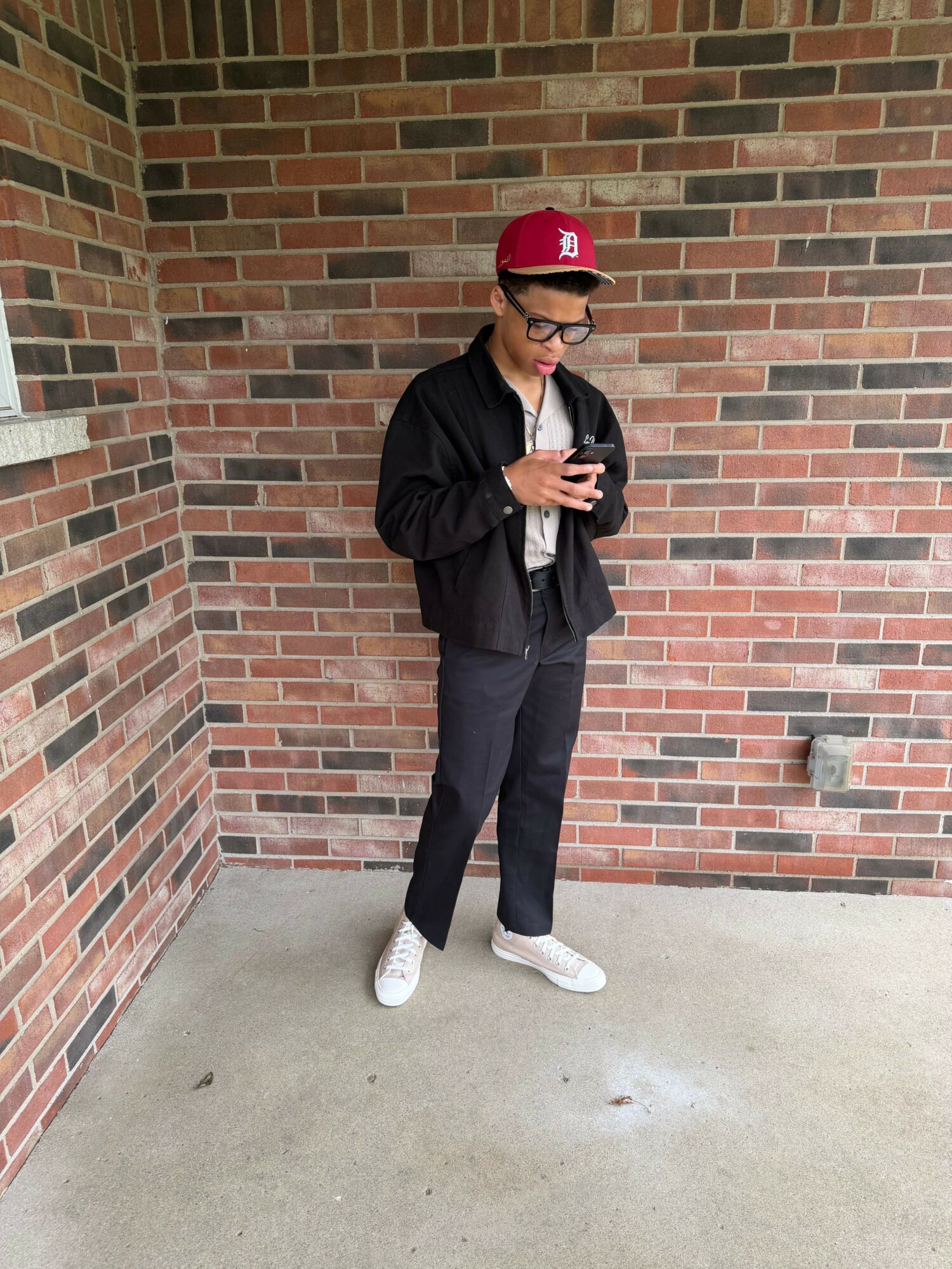 Person standing against a brick wall, wearing a red cap, black jacket, black pants, and white shoes, holding a phone.