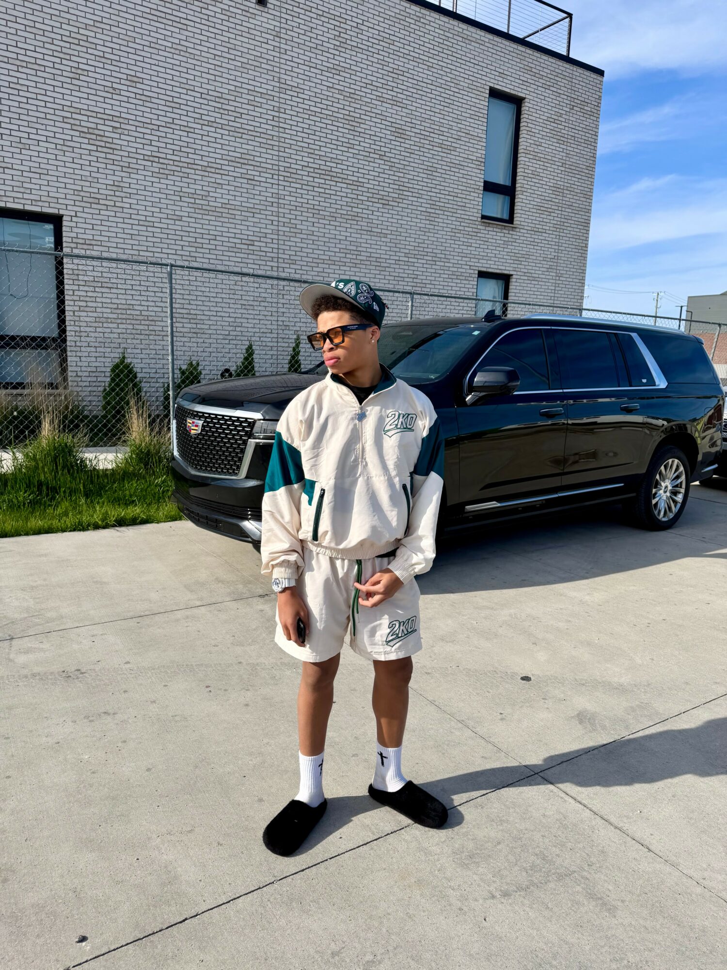A boy stands outdoors in front of a black car and a modern building, wearing a cap, glasses, and casual clothing.