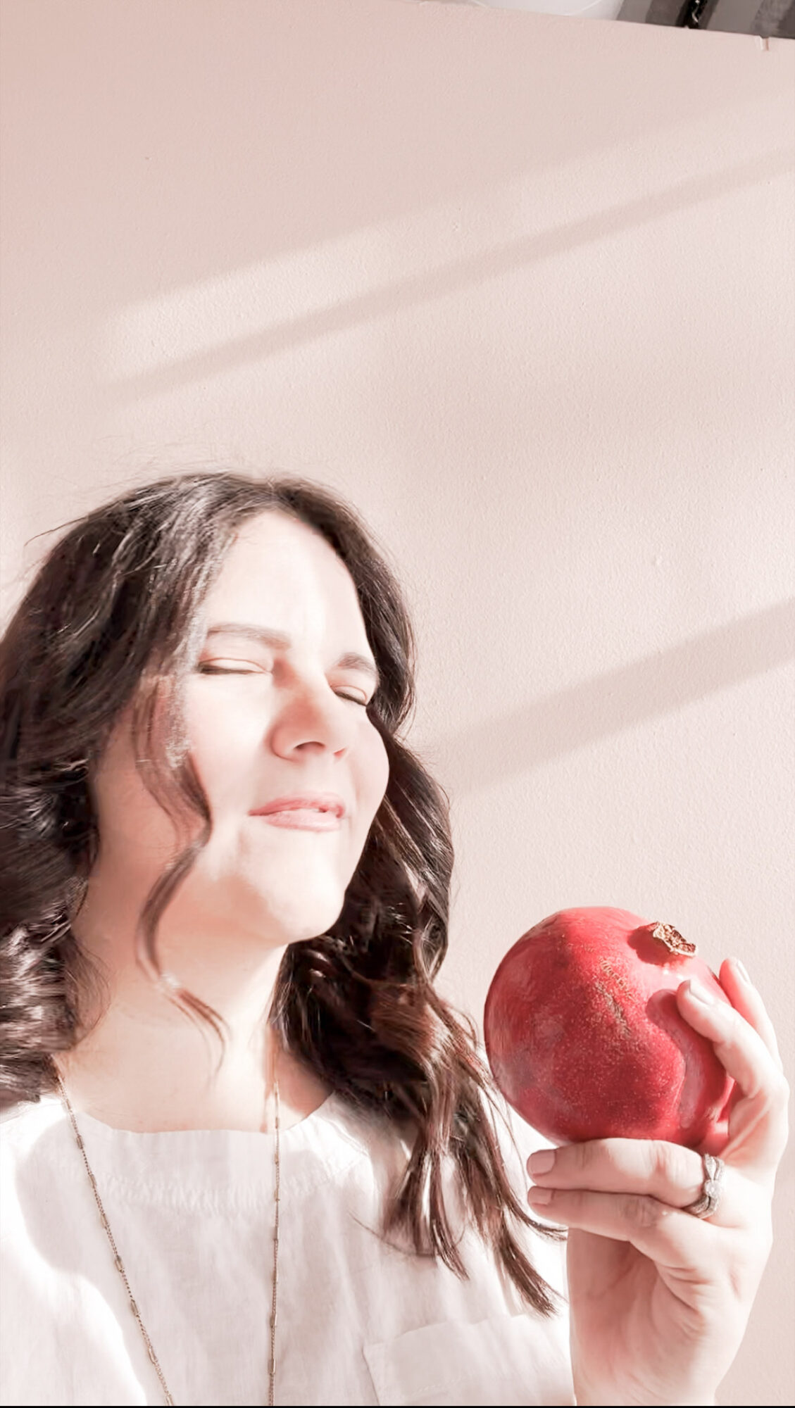 Woman with long hair holding a red apple, eyes closed, in bright light with shadows on wall