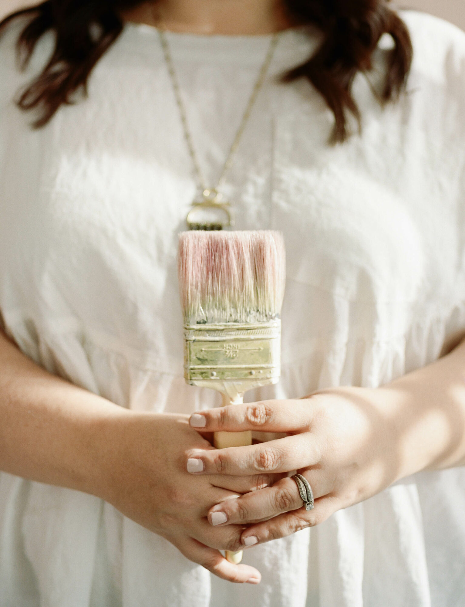 Person wearing a white dress holding a large paintbrush pendant necklace with both hands.