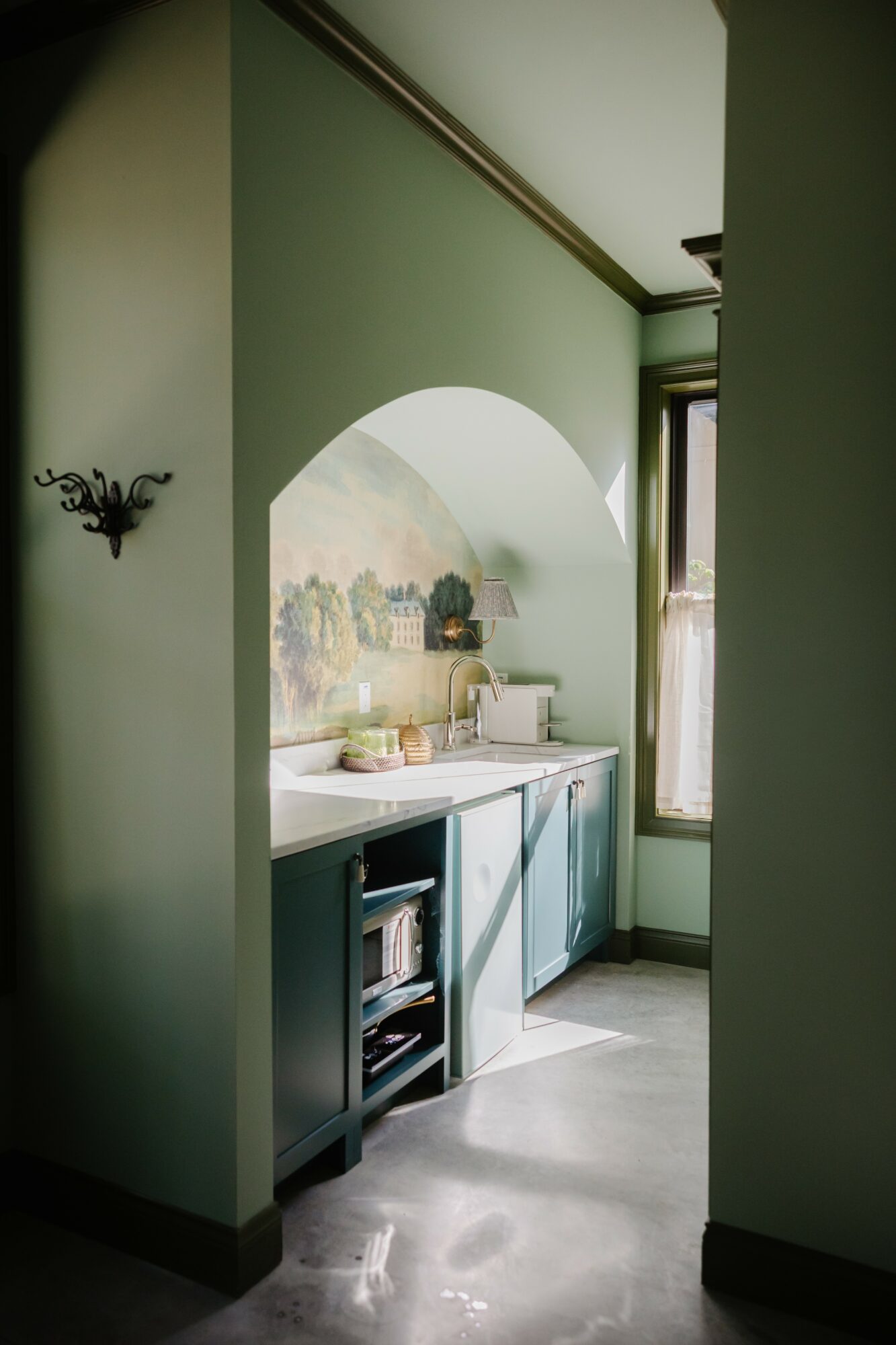 View through an archway into a small kitchen with green walls, a window, and a countertop with various items.