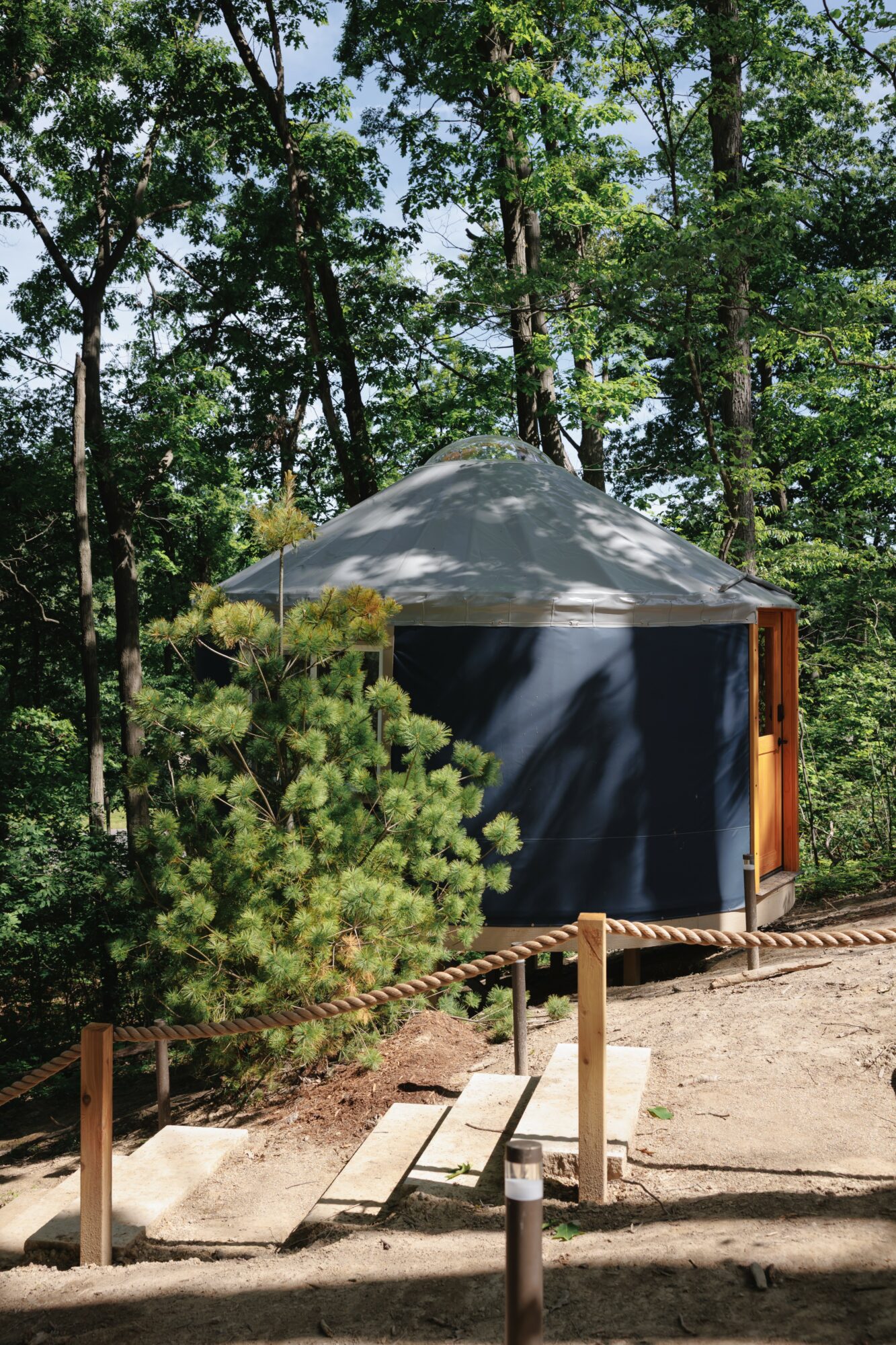 Small outdoor shelter with a gray roof, surrounded by trees and greenery, on a dirt ground with a wooden railing.