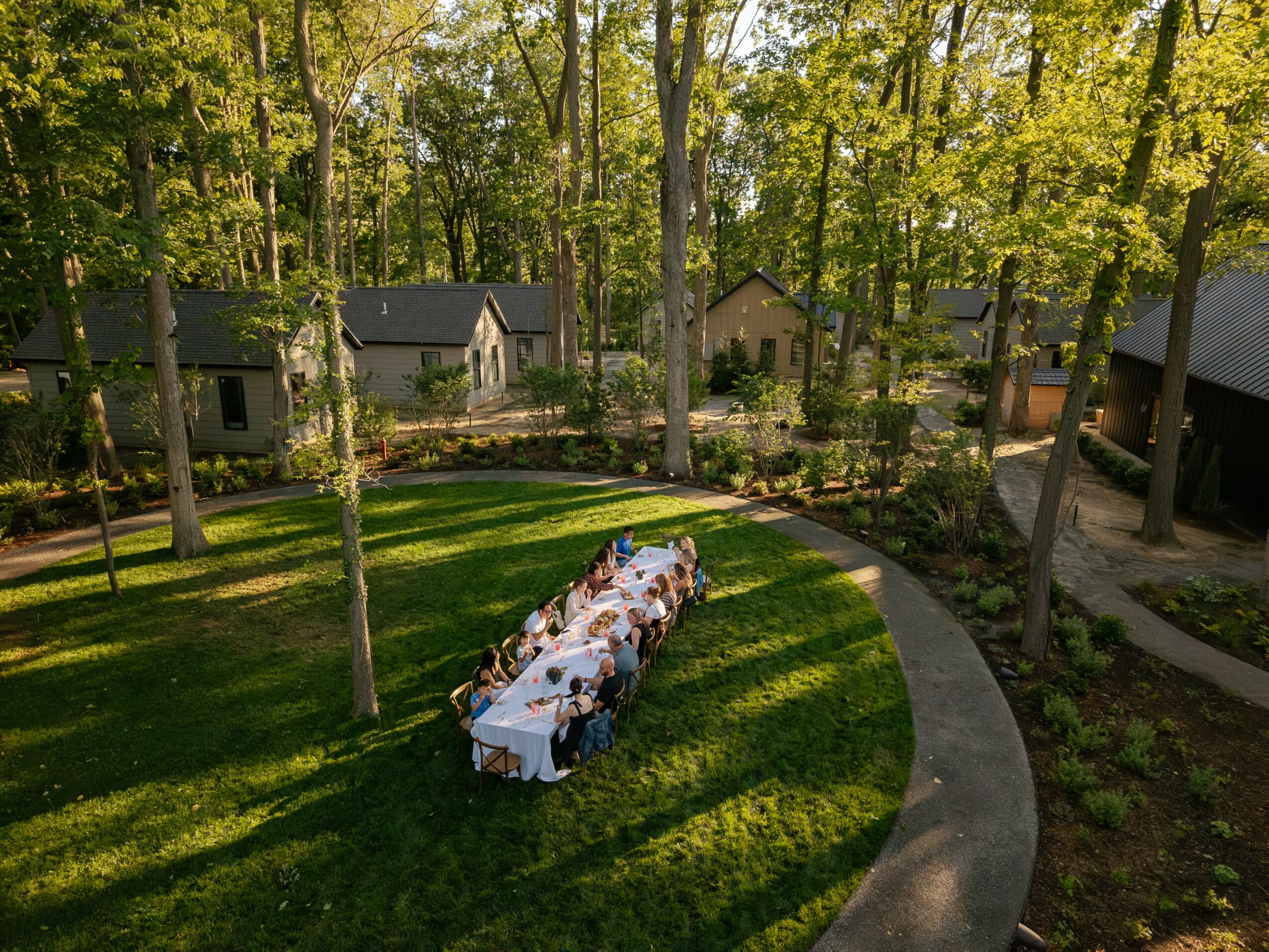 People sitting at a long table outdoors on a grassy area surrounded by trees and houses, with sunlight casting shadows.