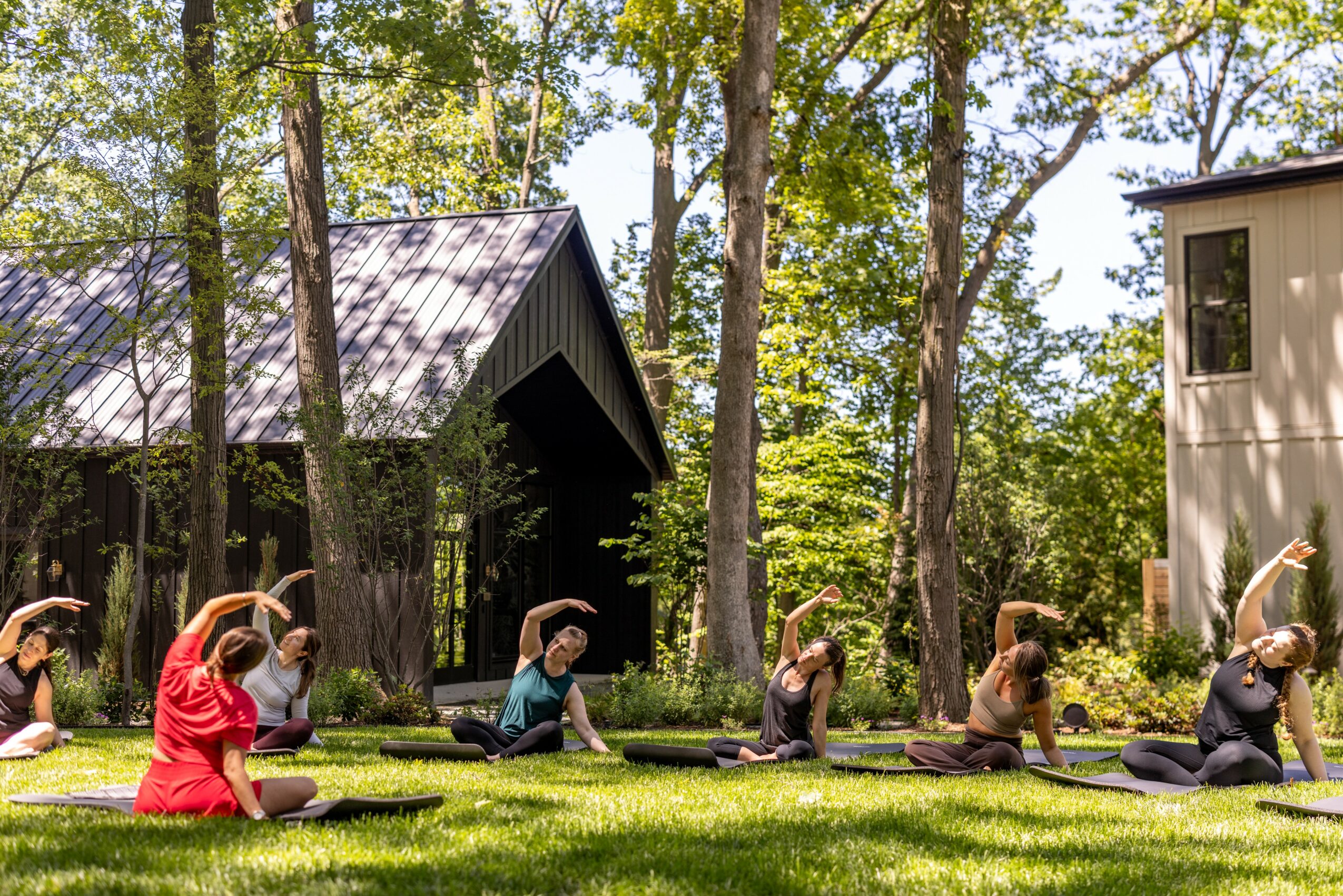 Group of people practicing yoga outdoors among trees and buildings, sitting on mats with arms raised overhead.