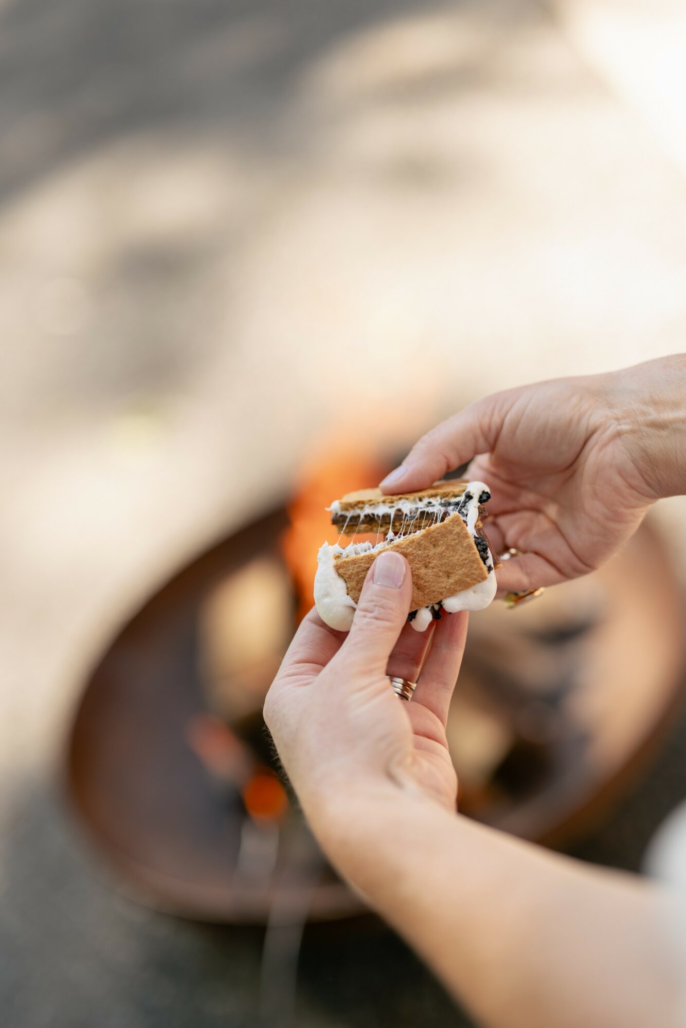 Person holding a piece of bread with melted chocolate, near a fire pit outdoors.